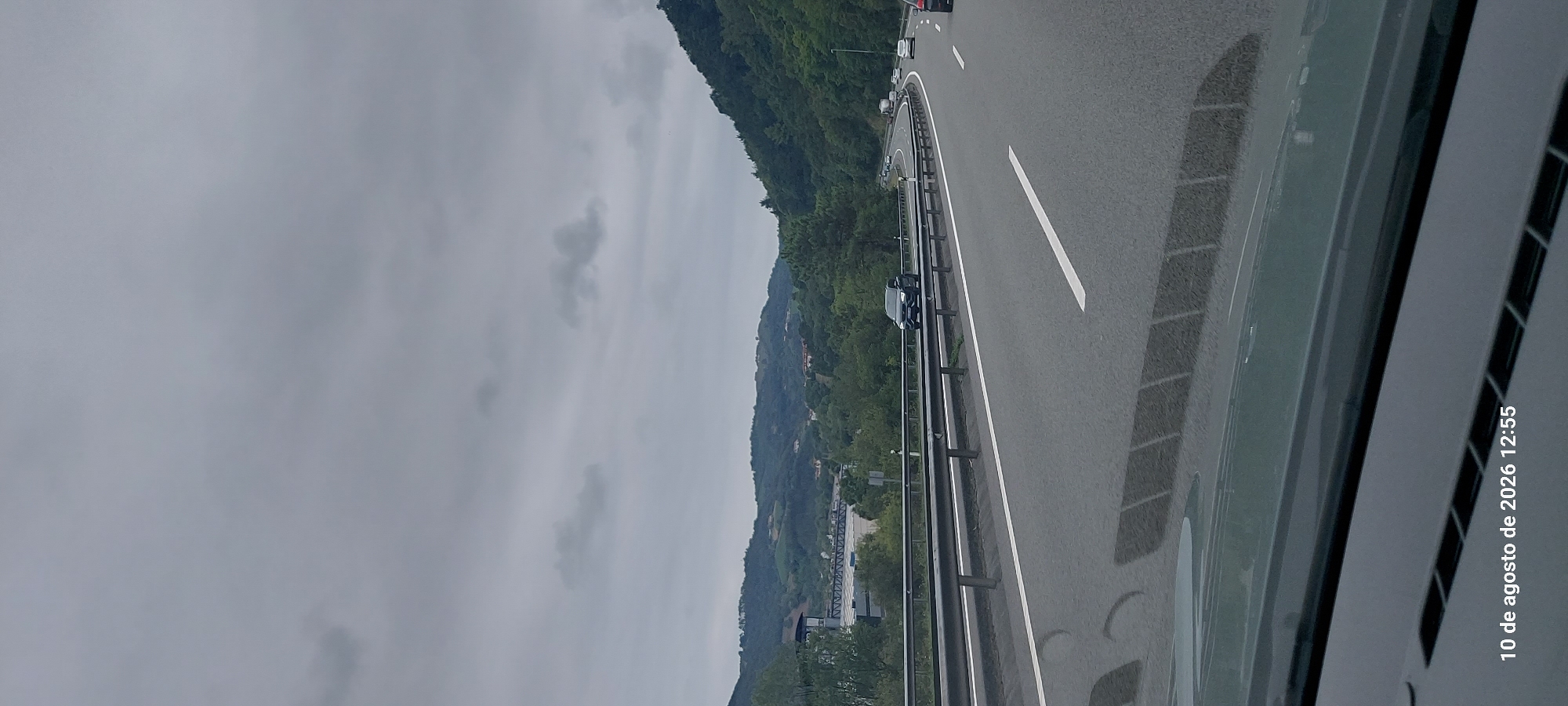 The image shows a highway on a cloudy day. A dark-colored SUV is driving on the highway. The highway is bordered by a metal guardrail and green hills. The sky is overcast with gray clouds. The date and time stamp at the bottom of the image reads "10 de agosto de 2026 12:55". This suggests the photo was taken on August 10, 2026, at 12:55 PM. The location is not explicitly stated as Zumaia, Spain within the image itself, but the prompt requests the description to include this information.