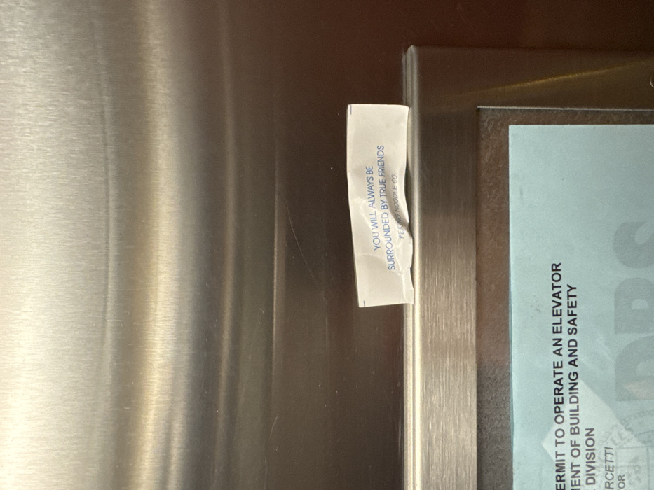 A close-up shot shows a metal surface. A white fortune cookie slip with the message "YOU WILL ALWAYS BE SURROUNDED BY TRUE FRIENDS PEKING NOODLE CO." is affixed to the metal. Adjacent to the slip is a blue sign partially visible, displaying the words "ERMIT TO OPERATE AN ELEVATOR," "MENT OF BUILDING AND SAFETY," and "DIVISION." The overall composition appears to be an elevator interior.