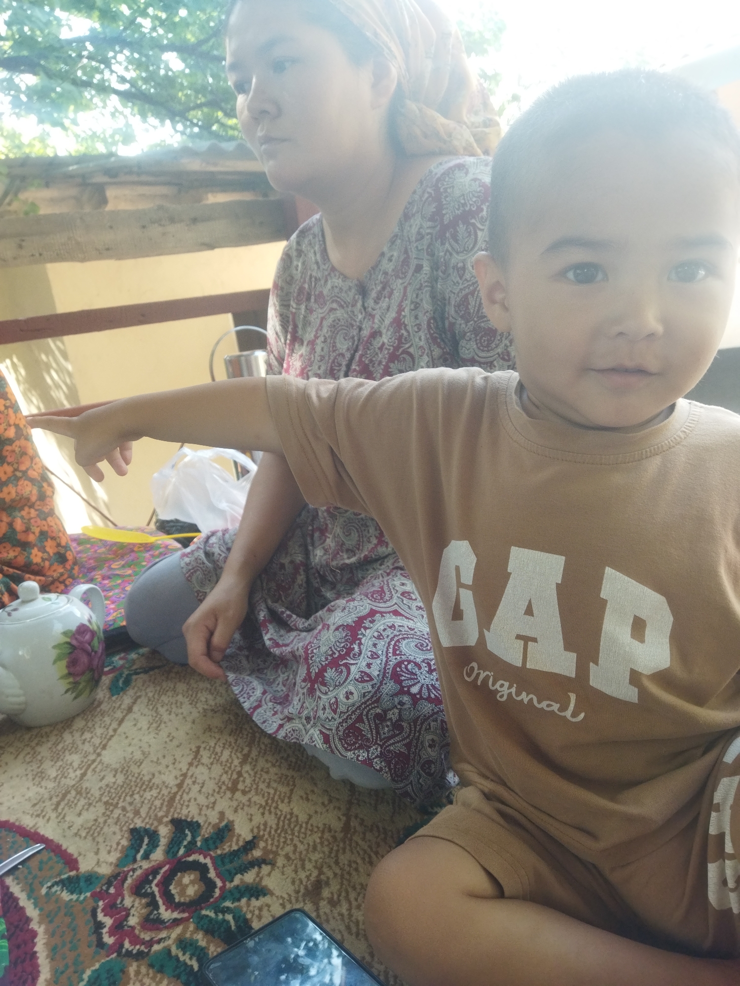 An outdoor scene in Mailuu-Suu City, Kyrgyzstan, features two individuals. In the foreground, a young boy wearing a tan t-shirt with "GAP Original" printed on it is seated. His right arm is extended, pointing towards the left. Beside him, a woman is seated, wearing a patterned headscarf and a dress with a maroon and white paisley print. She is looking to her left.
In the middle ground, a white teapot with a floral design sits on a patterned rug. A yellow utensil is visible nearby. A white plastic bag is also present. The background shows a yellow wall and some overhead foliage. The ground is covered with a carpet that has an intricate floral pattern in shades of beige, brown, green, and red. A mobile phone is partially visible at the bottom right of the frame.