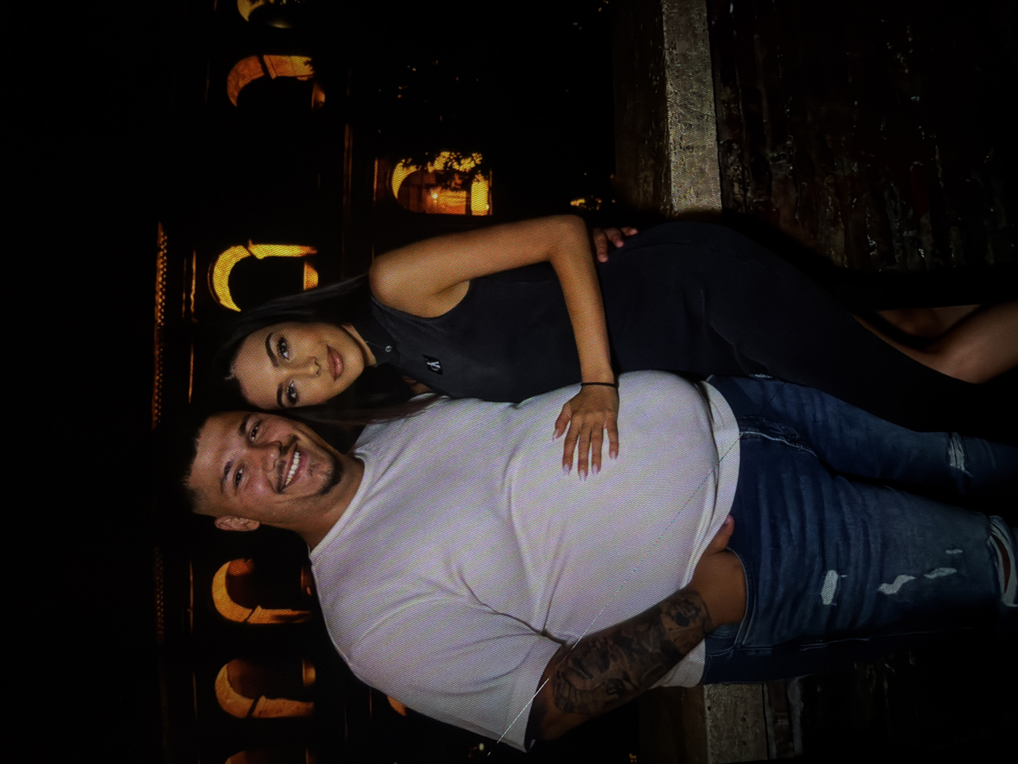 A medium shot captures a man and a woman posing for a photograph outdoors at night, with a dimly lit architectural structure featuring arched windows in the background. The man, positioned on the left, wears a light-colored t-shirt and ripped blue jeans, with tattoos visible on his left arm. He smiles warmly at the camera, with his right hand in his pocket and his left arm around the woman. The woman, on the right, is dressed in a dark, sleeveless dress and has her right arm around the man's waist. She looks towards the camera with a subtle smile. The scene is set against a dark backdrop, with the warm glow from the arched windows illuminating the couple and the immediate surroundings.