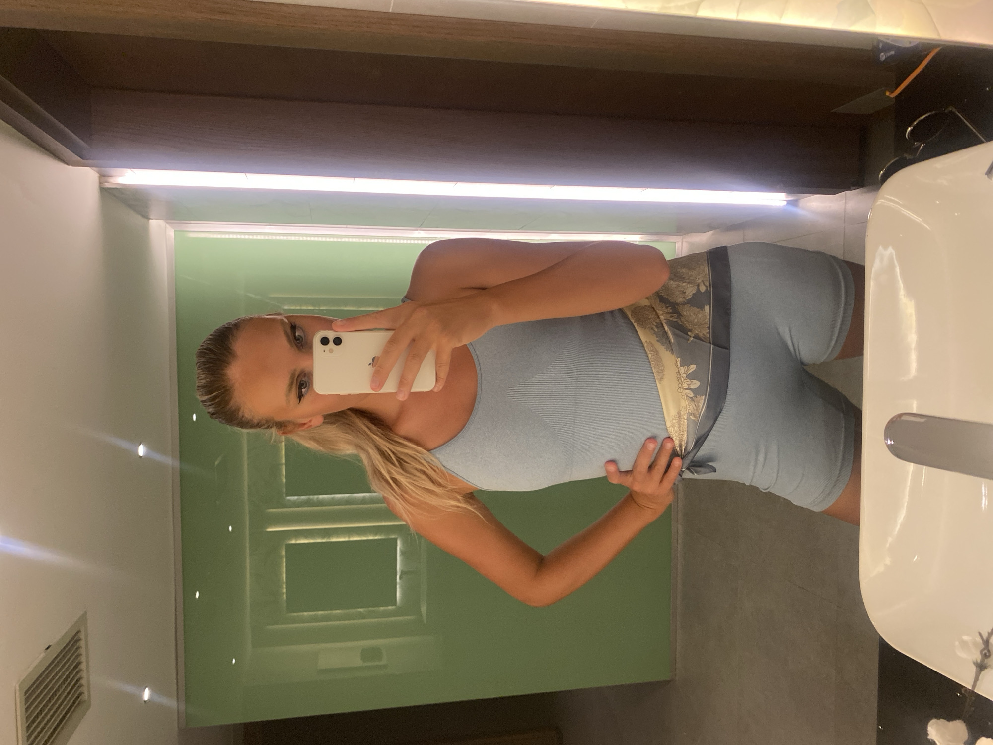 A woman with blonde hair pulled back in a ponytail takes a mirror selfie in what appears to be a modern hotel bathroom. She is wearing a light blue ribbed tank top and matching short shorts, cinched at the waist with a patterned silk scarf. Her left hand is on her hip, and she holds a white smartphone in her right hand to capture the reflection. The bathroom features mint green walls, a large mirror, and bright vertical lighting. The foreground includes a white sink basin. The scene is located in Aksu, Turkey.