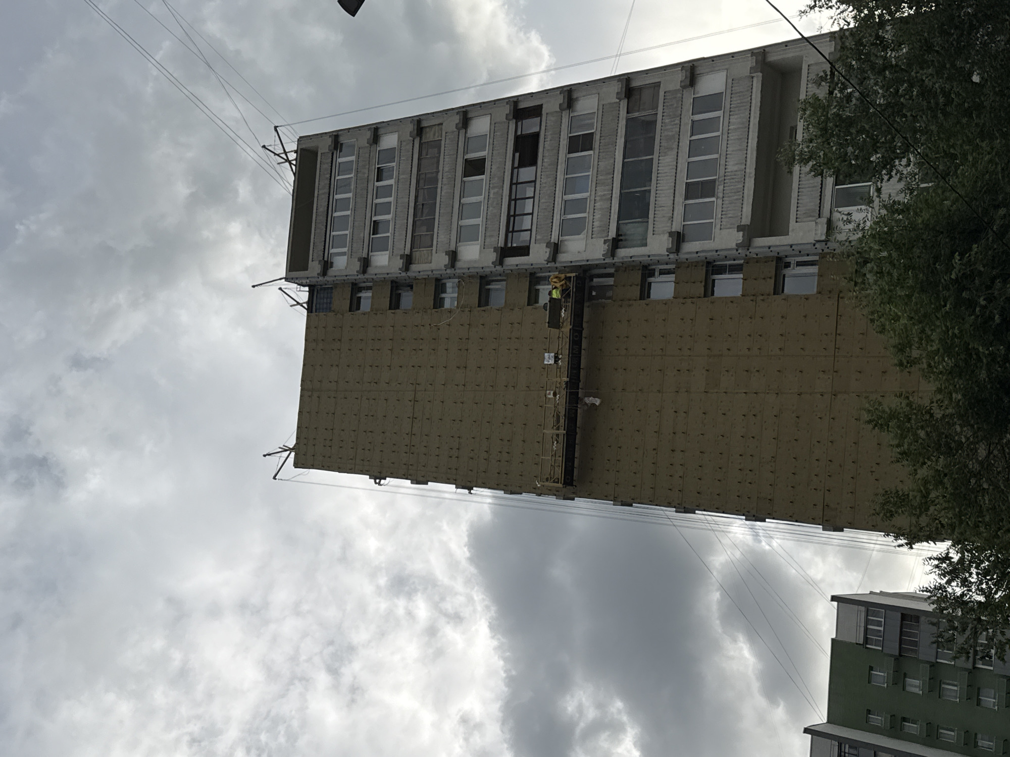 This is an upward-facing, wide-angle shot of a building undergoing exterior renovation under a cloudy sky. The primary subject is a tall, rectangular building with a section of its facade covered in beige insulation panels. Above these panels, the original building facade is visible, featuring rows of rectangular windows with grey frames. Some of these windows are open, revealing dark interiors or ladders.
At the mid-section of the building, a small construction platform or hoist is attached to the facade. A lone construction worker in a yellow vest is visible on this platform, suggesting active renovation work is in progress. The building appears to be of a somewhat dated architectural style, possibly from the Soviet era, given its modular window design.
Further down and to the right, a glimpse of another building with a green facade and multiple windows is visible, hinting at a dense urban environment. Overhead power lines crisscross the sky, suggesting an urban or suburban setting. A leafy tree on the right partially obstructs the view of the renovation site.
The weather appears overcast and possibly humid, with dramatic cloud formations dominating the sky. The lighting is diffused, indicating it is daytime, likely late afternoon or early morning given the soft light. The overall scene suggests a typical day of construction work in a Russian city, likely Moscow as per the location context. No visible text is present in the image. The mood is neutral, focused on the construction activity.
