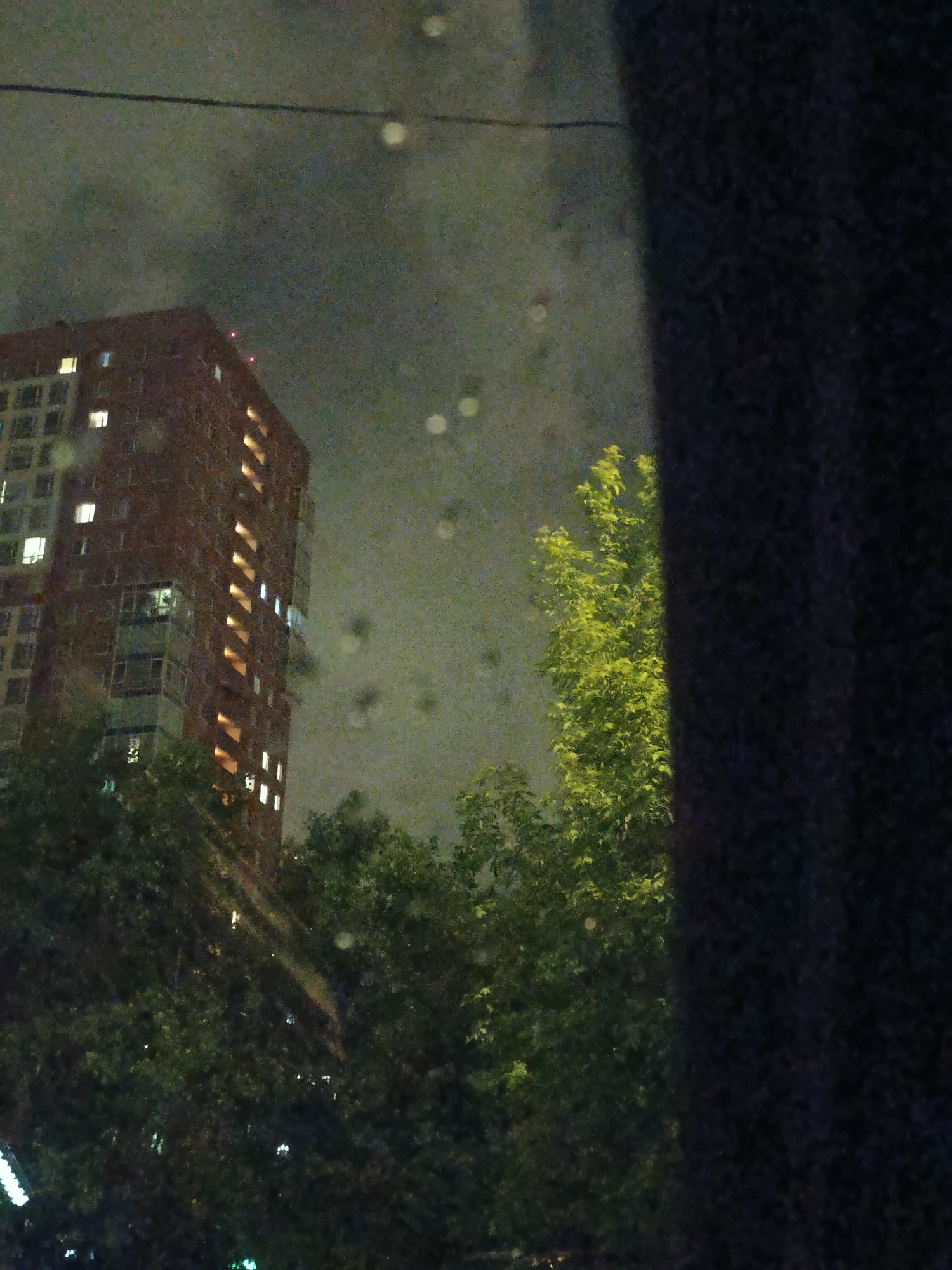 A night scene shows a tall, brick apartment building with lighted windows, partially obscured by trees. To the right, a dark vertical element, possibly part of the architecture or a curtain, frames the view. The location is likely Moscow, Russia based on the style of the building. The sky is overcast. There are rain droplets on the window in the foreground.