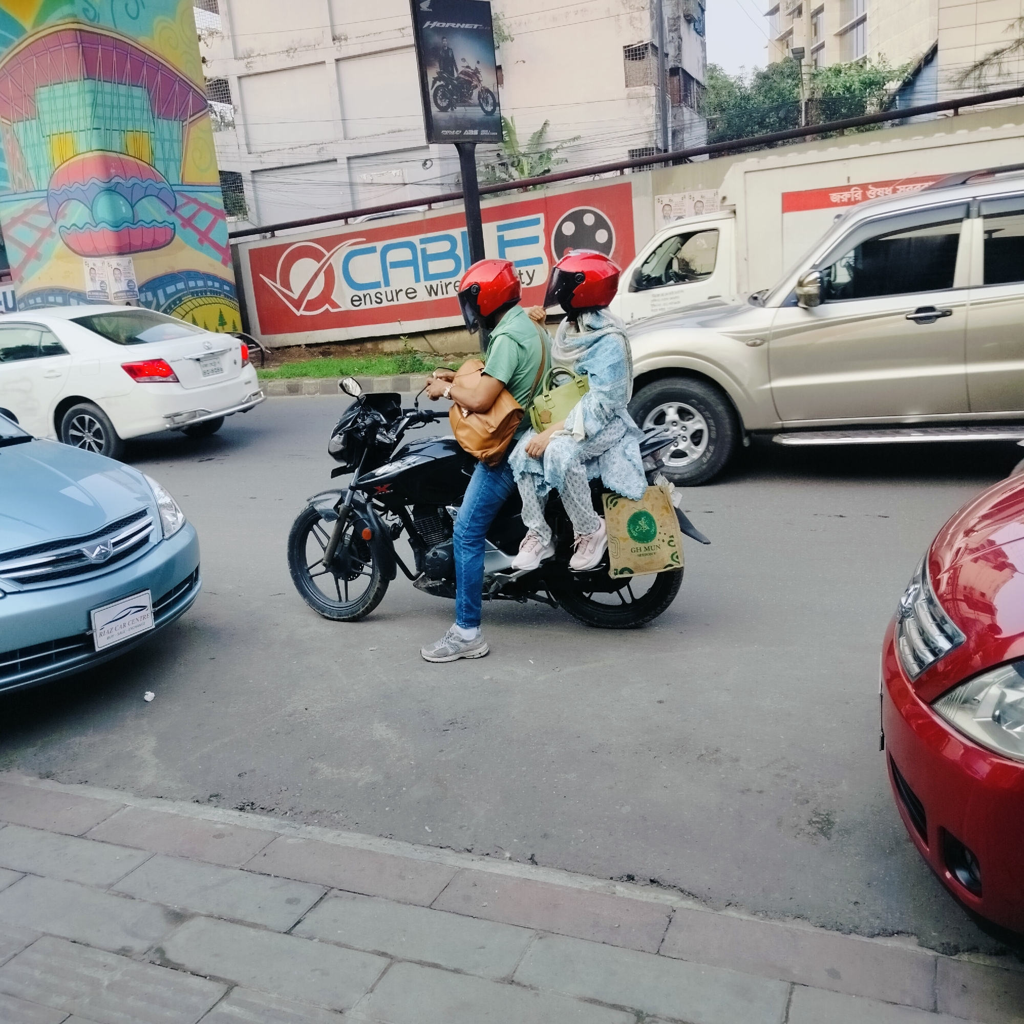 The image captures a street scene in Dhaka, Bangladesh. A man and a woman, both wearing matching red helmets, are on a black motorcycle stopped at a street corner. The man, dressed in a green shirt and blue jeans, is seated on the motorcycle, while the woman, wearing a blue patterned dress and a light-colored headscarf, sits behind him. The man carries a brown satchel, and the woman holds a green purse and a shopping bag. Several cars are visible around them: a light blue sedan, a white sedan, and a tan SUV. The front of a red car is on the right edge of the photo.
Behind the motorcycle and cars, a sign advertises "CABLE ensure wire ty." Above the sign, a billboard displays an image of a Honda Hornet motorcycle. A partially visible building with a colorful mural adorns the side, and another building is in the background.
Text on the blue sedan reads "RIAZ CAR CENTRE," which helps to identify the location in Dhaka. The street appears busy with traffic.