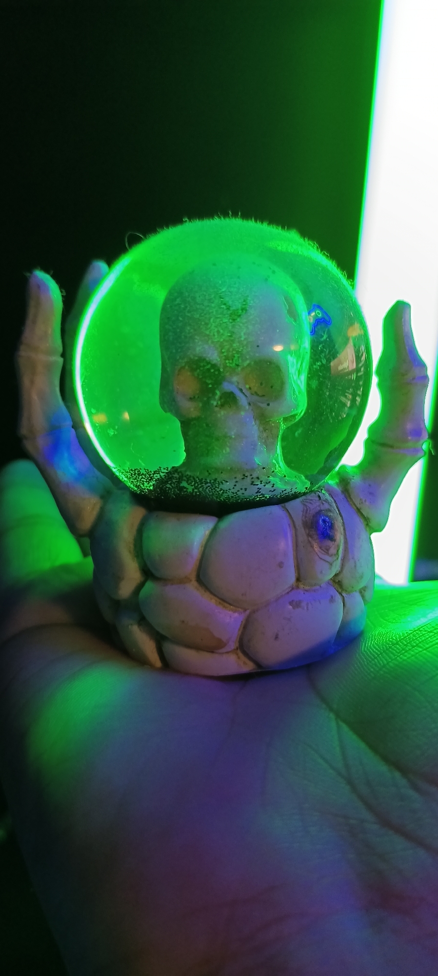 A close-up, handheld shot features a novelty skull figurine resting on a person's palm, illuminated by vibrant green and purple lighting. The figurine, made of a pale, textured material, depicts a skull encased in a clear, green sphere. The base of the figurine is designed to resemble segmented, bony structures, possibly ribs or armor, and is supported by what appear to be skeletal hands. The person's palm, visible at the bottom of the frame, displays distinct lines and is cast in shadows and reflections from the colored lights. The background is dark with a bright, vertical light source on the right side of the image, creating a strong contrast. The overall mood is atmospheric and somewhat eerie, amplified by the intense lighting and the macabre subject matter. There is no indication of the city of Wels, Austria, in the image or any information that would suggest this location.