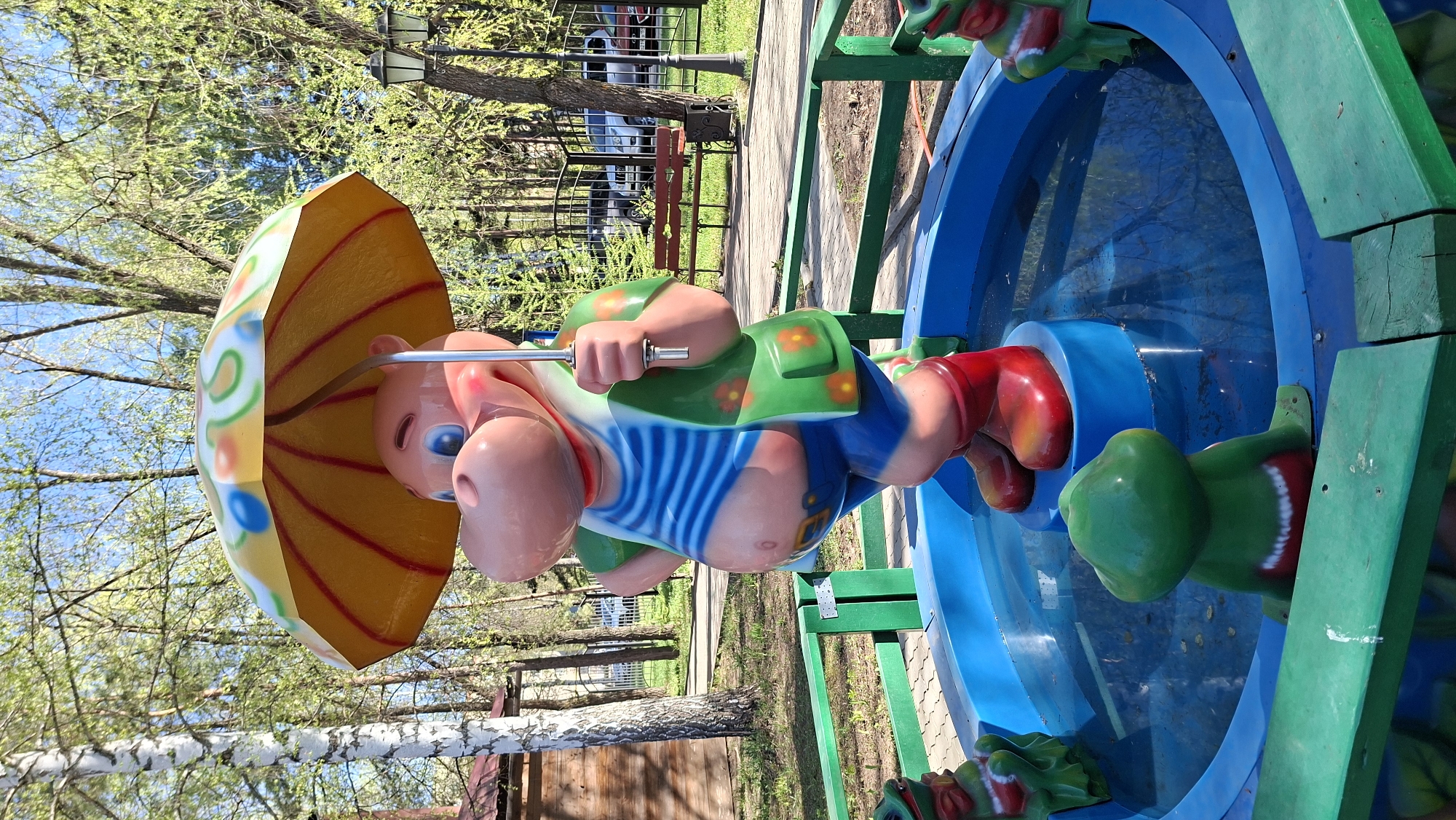 The image shows a children's carousel in what appears to be a park setting. The carousel features colorful characters, one of which is a pink figure with a striped shirt and a yellow umbrella. The structure is made of blue and green painted metal, set against a backdrop of trees and a glimpse of what could be park benches and street lamps. Based on the location, this likely is a park or amusement area in Chaikovsky, Russia.