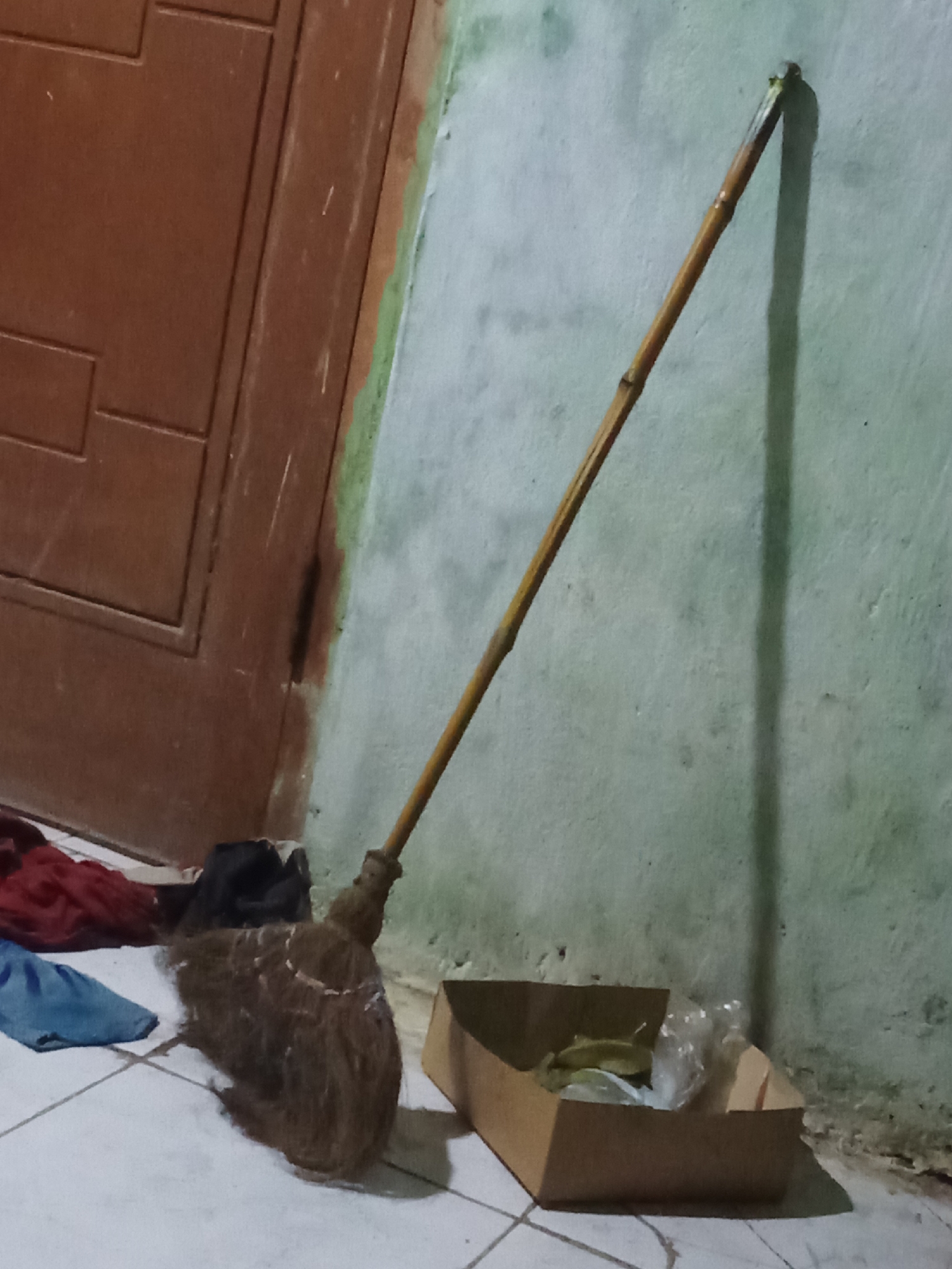A broom with a bamboo handle leans against a textured, pale green wall. The broom's bristles are made of natural fibers and appear worn. To the left of the broom, a brown wooden door with recessed rectangular panels is visible. In the foreground, a few pieces of folded fabric, one red and one blue, lie on a tiled floor. To the right of the broom, a small, opened cardboard box contains some indistinct items, possibly trash or discarded materials, some wrapped in plastic. The lighting suggests an indoor environment, possibly a domestic or storage area, with a shadow cast by the broom on the wall. The scene is devoid of any individuals. The overall impression is one of a utilitarian space.