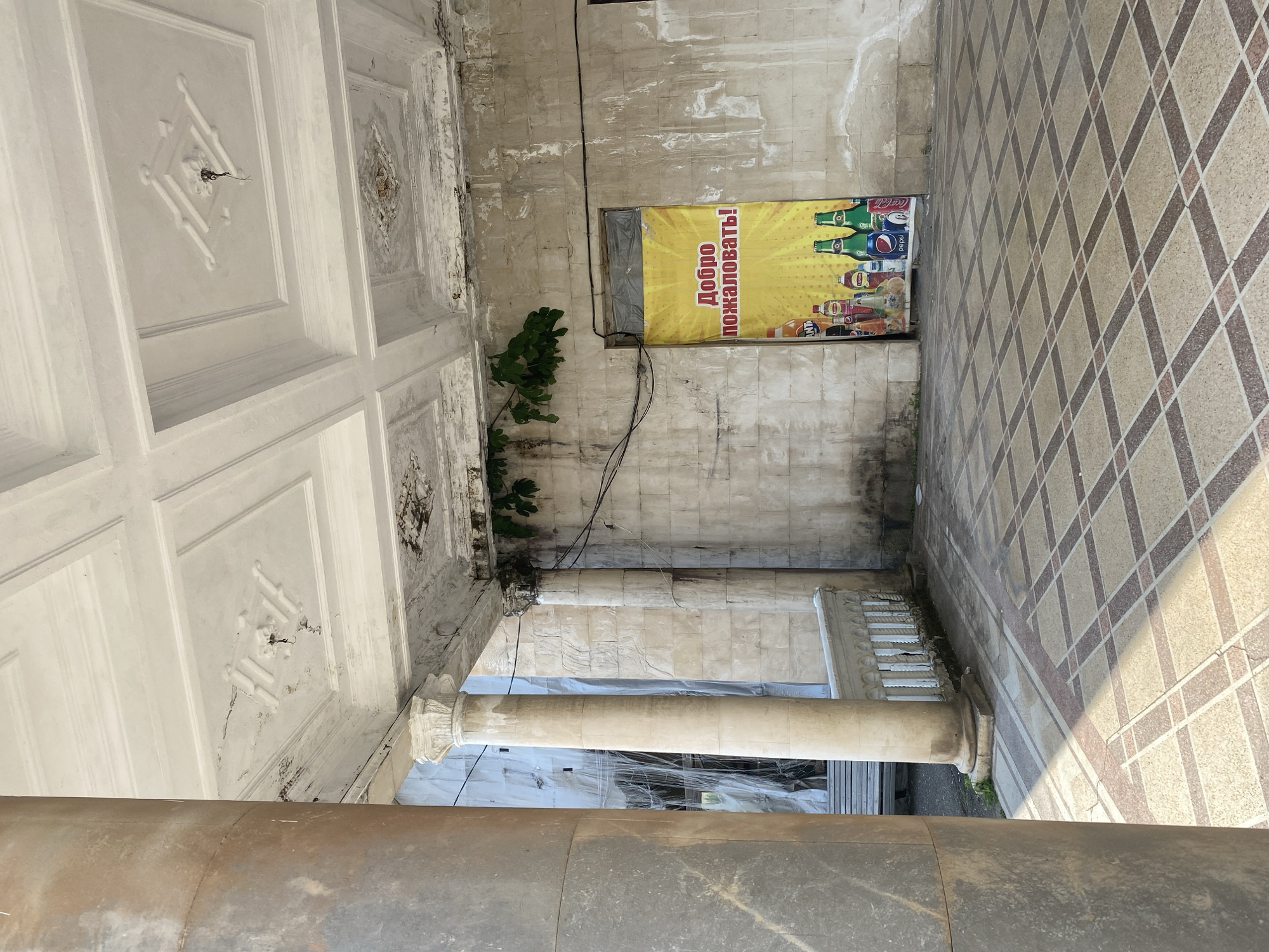 The image depicts an exterior view of a building with architectural elements that suggest a historical or possibly abandoned establishment in Psakhara, Abkhazia. The foreground features a patterned tiled floor, with sunlight casting distinct shadows. To the left, a large, weathered stone column stands prominently. Beyond this column, another lighter-colored column supports an ornate, aged ceiling with decorative moldings and signs of water damage and peeling paint. The wall behind the columns is constructed of large, rough-hewn stone blocks. A decorative white railing runs along a raised area. In the center-right of the image, a doorway is covered by a large yellow banner displaying the Russian phrase "Добро пожаловать!" (Welcome!) in red and white text, superimposed over a bright yellow sunburst graphic and images of various beverage bottles. Some green foliage is visible growing from the top of the wall near the banner. The overall impression is one of a once grand building now showing significant signs of disrepair and weathering, with the banner indicating a potential commercial use or entrance to a business. In the background, through an opening to the left, construction materials or tarpaulin are visible, suggesting ongoing or suspended renovation work.