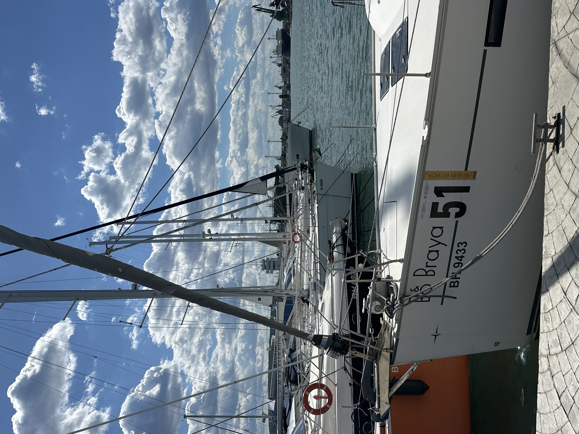 The image shows several yachts docked in a harbor. The foreground features the stern of a large white yacht named "Braya," displaying the number "51" and the registration "BH 9433." It also has a sticker related to the "Aegean Regatta" for 2025. Adjacent to this yacht are other yachts with masts and rigging visible against a backdrop of a blue sky filled with puffy white clouds. In the distance, the city of Varna, Bulgaria, is partially visible, including what appears to be an industrial area with cranes along the waterfront. The yacht is docked alongside a paved walkway. The name of "BLACK SEA YACHTING" is visible on another boat along with the registration "Vn 8666."