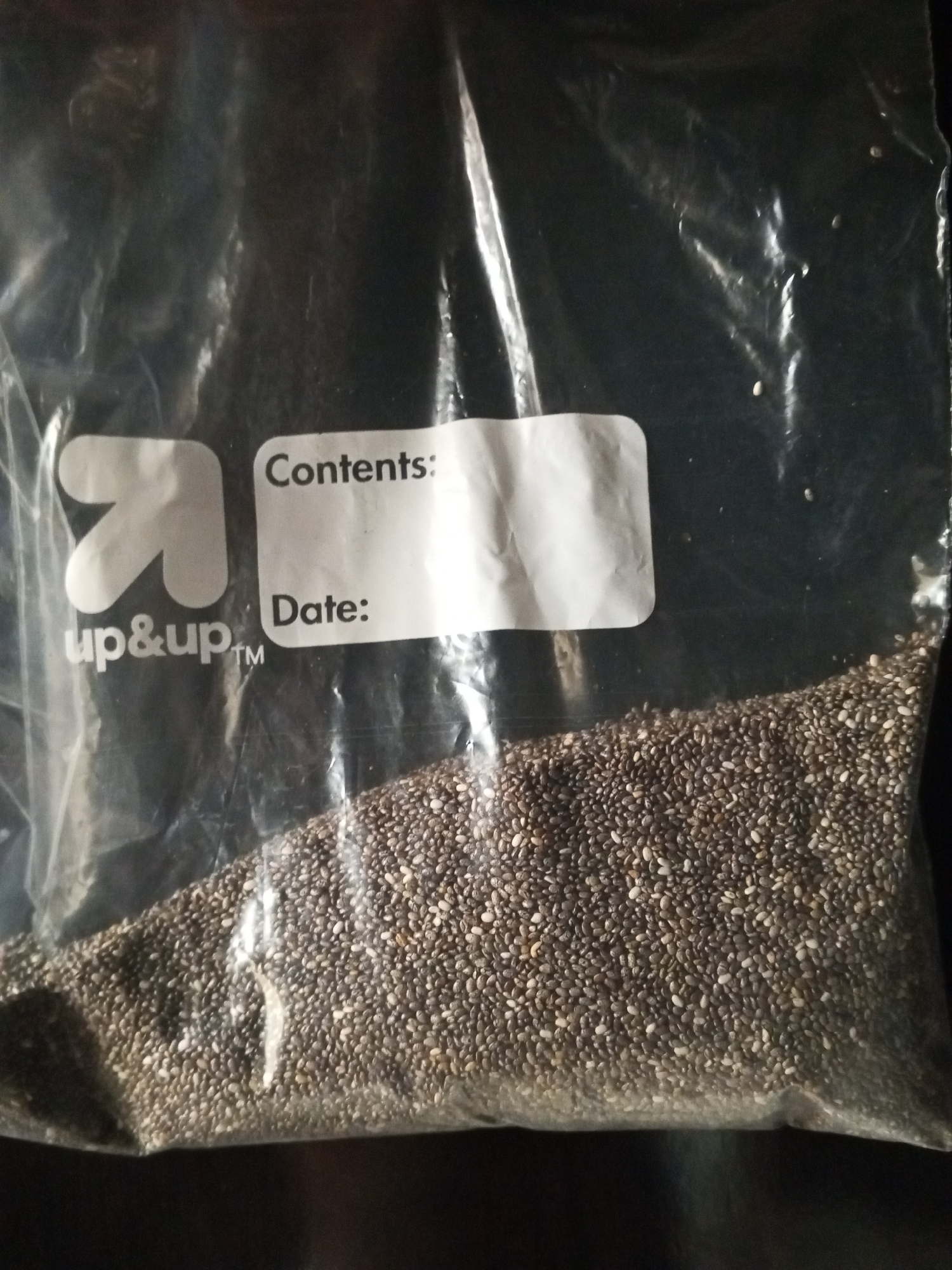 The image shows a clear plastic bag filled with chia seeds. The bag has a white label with the "up&up" logo and text that reads "Contents:" and "Date:". The bag is positioned on a dark, textured surface. There are no people visible in the image. The lighting suggests an indoor setting, possibly a kitchen or pantry. There is no indication of the location being Ashata, Nigeria.