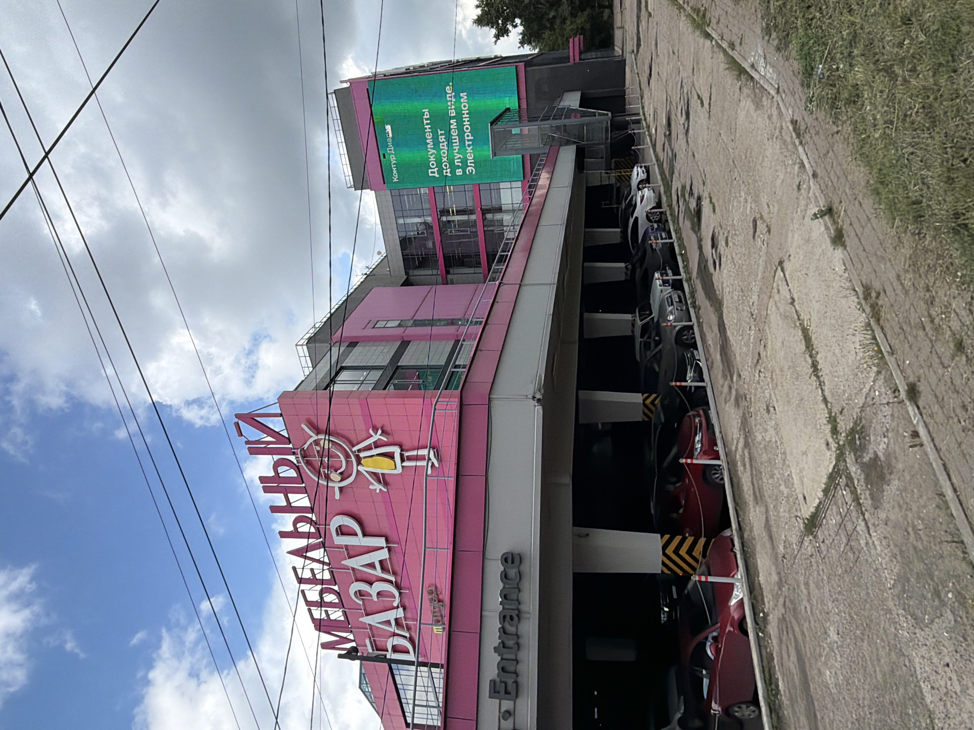 The image shows the exterior of a large building, likely a shopping center, with "МЕБЕЛЬНЫЙ БАЗАР" (Furniture Bazaar) prominently displayed on its pink facade. The building has modern architecture with large windows and a digital billboard on the right side displaying Russian text that translates to "Documents arrive in the best form. Electronic." Below the main facade is a covered parking area where several cars are parked. The area around the building appears to be an urban environment with a cloudy sky overhead and some trees visible in the distance. The building's location is identified as Nizhny Novgorod, Russia, based on information provided. There are no visible people in the image.