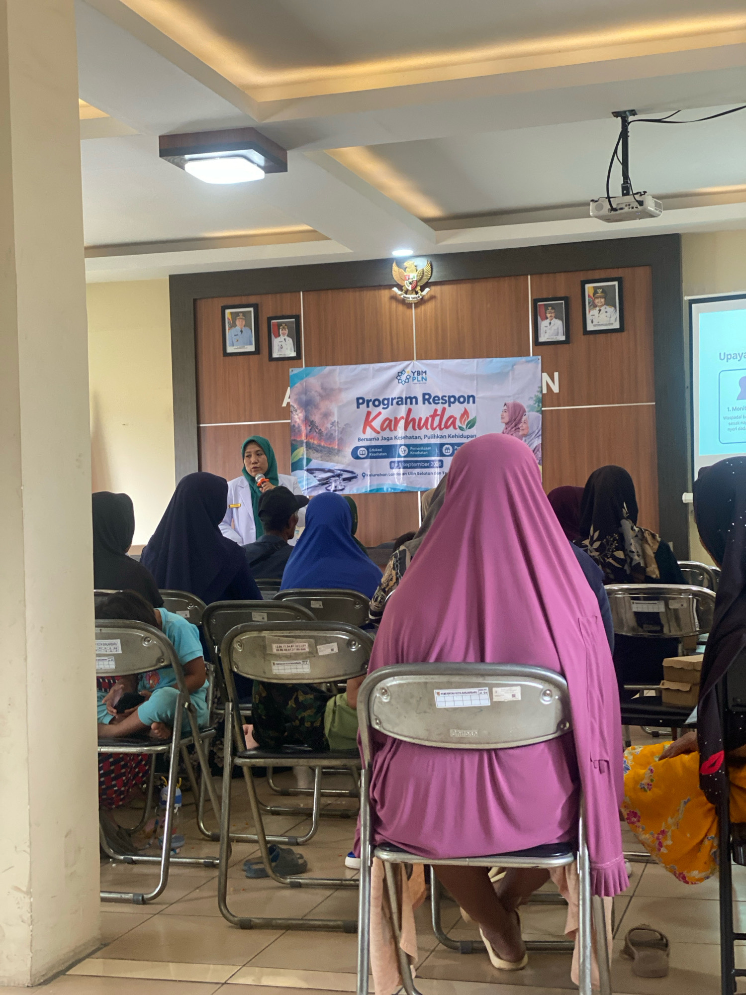 An indoor scene, likely a meeting room in Banjarbaru, Indonesia, shows a woman in a white lab coat and green hijab speaking into a microphone at the front of the room. She is standing in front of a banner that reads "PROGRAM RESPON KARHUTLA Bersama Jaga Kesehatan, Pulihkan Kehidupan" with the PLN and YBM logos. A projector is mounted on the ceiling. Several people, mostly women wearing hijabs, are seated in chairs facing the speaker. One person in the foreground wears a bright pink hijab. The room has a tiled floor and is lit by overhead fluorescent lights. Pictures of officials are displayed on the wooden-paneled wall behind the banner. The overall atmosphere suggests an educational or community event.