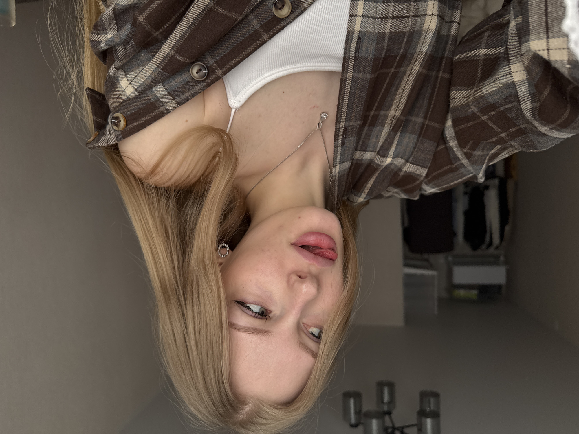 A young woman with blonde hair and blue eyes is posing for a photo. She is wearing a white tank top under a brown and grey plaid flannel shirt. She has a silver necklace and earrings. Her tongue is slightly sticking out. The background is a plain wall. The photo is rotated sideways. The location is not identifiable as Perm, Russia based on the image.