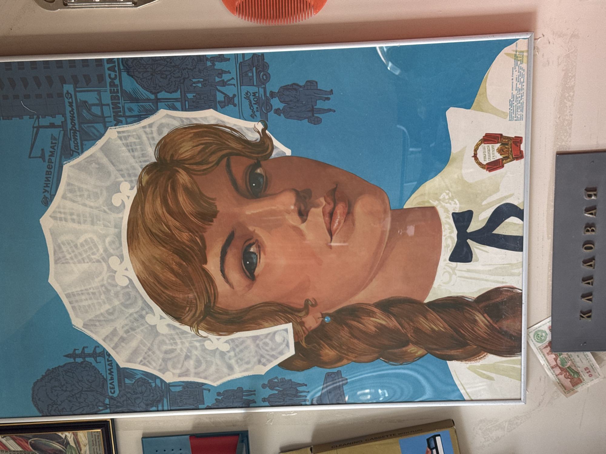 A framed Soviet-era commercial poster, oriented vertically, dominates the image, depicting a woman's face and upper torso against a blue background. The woman, with light brown hair styled in a braid and wearing a white lacy cap and a white dress with a black bow, gazes directly at the viewer with light-colored eyes and a serene expression.
The blue background of the poster features stylized silhouettes of urban and rural landscapes, including buildings, trees, pedestrians, and a delivery truck, suggesting various commercial settings. Visible Cyrillic text on the poster promotes different types of stores and services: "УНИВЕРМАГ" (Universal Store), "ГАСТРОНОМ" (Grocery Store), "УНИВЕРСАЛ" (Universal, likely part of a store name), "СЕЛЬМАГ" (Rural Store), and "ДОСТАВКА НА ДОМ" (Home Delivery). A small text block in the upper right corner indicates the artist "М. Стерников" and the year "1955 г." (1955), published by "ИЗДАТЕЛЬСТВО СОВЕТСКИЙ ХУДОЖНИК" (Soviet Artist Publishing House). An emblem near her right shoulder also contains Cyrillic text, possibly referencing an exhibition.
The poster is displayed indoors on a light-colored wall. Above it, part of a red plastic comb is visible. To the right, a dark gray sign with embossed Cyrillic letters "КЛАДОВАЯ" (Storeroom) is partially visible. Below this, a crumpled Soviet 5-ruble banknote is present. The overall scene suggests a collection of historical artifacts or items displayed in a private setting in Perm, Russia.