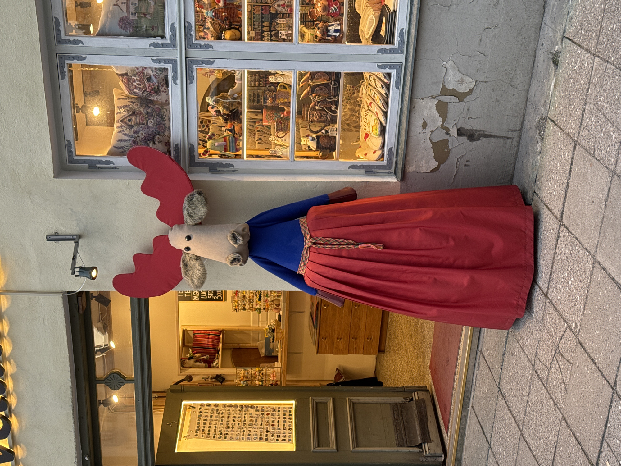 This is a full, eye-level, outdoor shot of a stuffed moose figure dressed in a red skirt and blue top, standing in front of a shop window in Tallinn, Estonia. The figure is the main subject, with its large red antlers prominent. The shop window displays various items, including mugs, figurines, and cushions. The shop entrance is open to the left, revealing a counter and more merchandise inside. The ground is paved with cobblestones, and the building facade shows signs of wear and tear. The lighting suggests it is late afternoon or early evening.