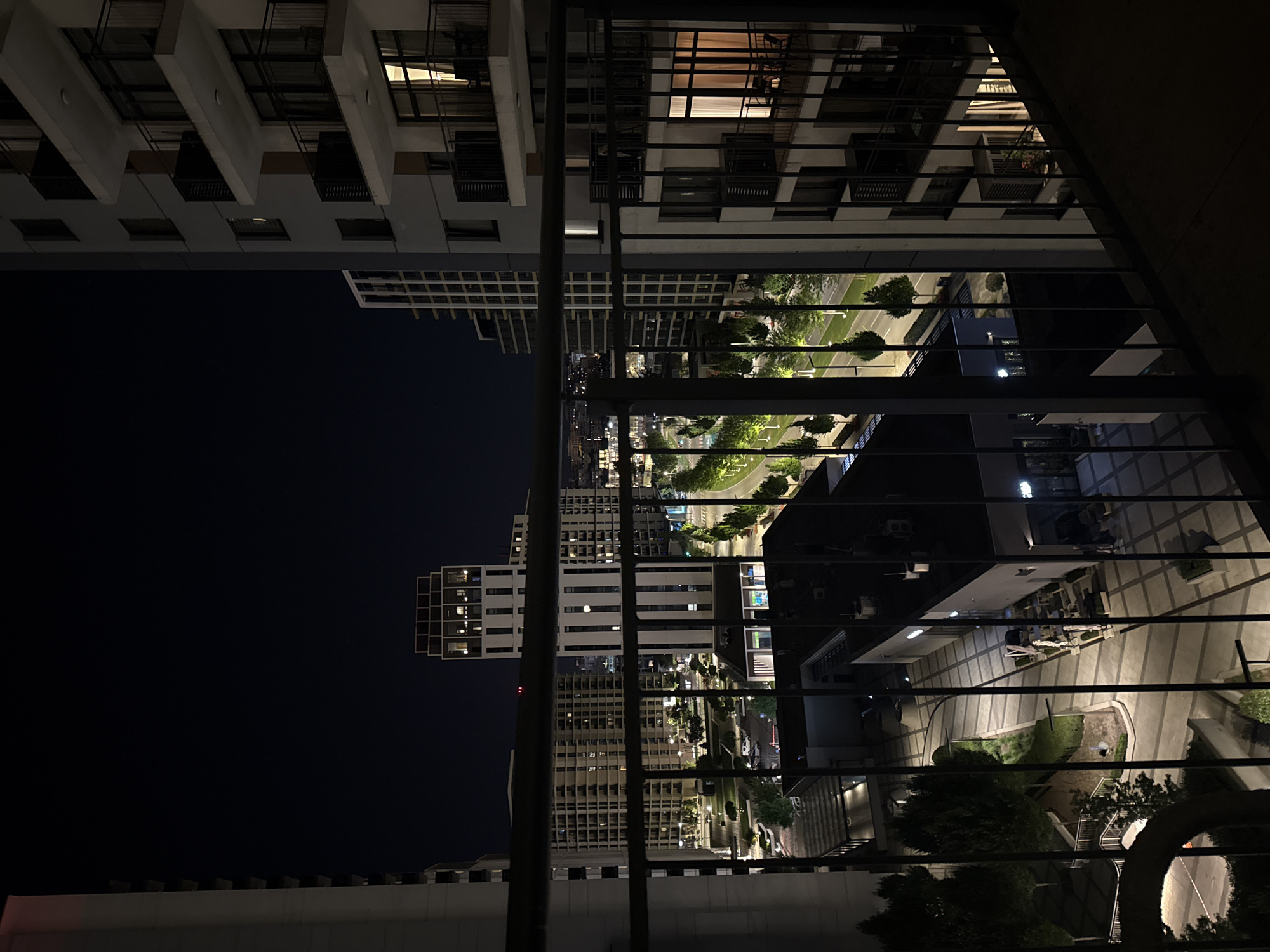 This is a nighttime outdoor photograph taken from a balcony overlooking a modern urban environment in Belgrade, Serbia. The foreground is dominated by a dark metal railing, characteristic of a balcony. Beyond the railing, a courtyard area is visible, illuminated by streetlights and lights from within buildings. The courtyard features paved walkways, scattered trees, and low-lying green areas. Several modern buildings are present, including apartment complexes and taller structures with many windows, some of which are lit. A road or pathway curves through the scene, lined with trees that are lit by artificial lights, casting green hues. The sky is dark and clear, indicating night. There are no visible people in the scene. The overall impression is of a well-lit, contemporary urban development at night.