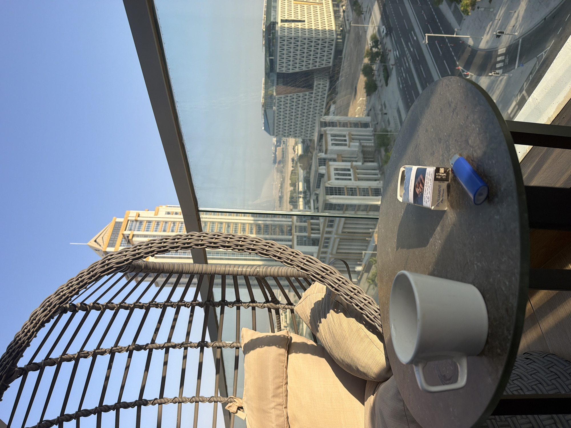 A low-angle, outdoor shot captures a balcony scene overlooking a city with modern architecture under a clear blue sky. In the foreground, a round, dark gray table holds a white mug, a pack of Parliament cigarettes, and a blue lighter. To the left, a woven hanging chair with beige cushions is partially visible. Beyond the table and chair, a cityscape unfolds, featuring tall buildings and a multi-lane road with sparse traffic. The lighting suggests it is daytime, with sunlight casting shadows from the chair onto the table. The overall impression is one of a quiet, elevated urban dwelling.