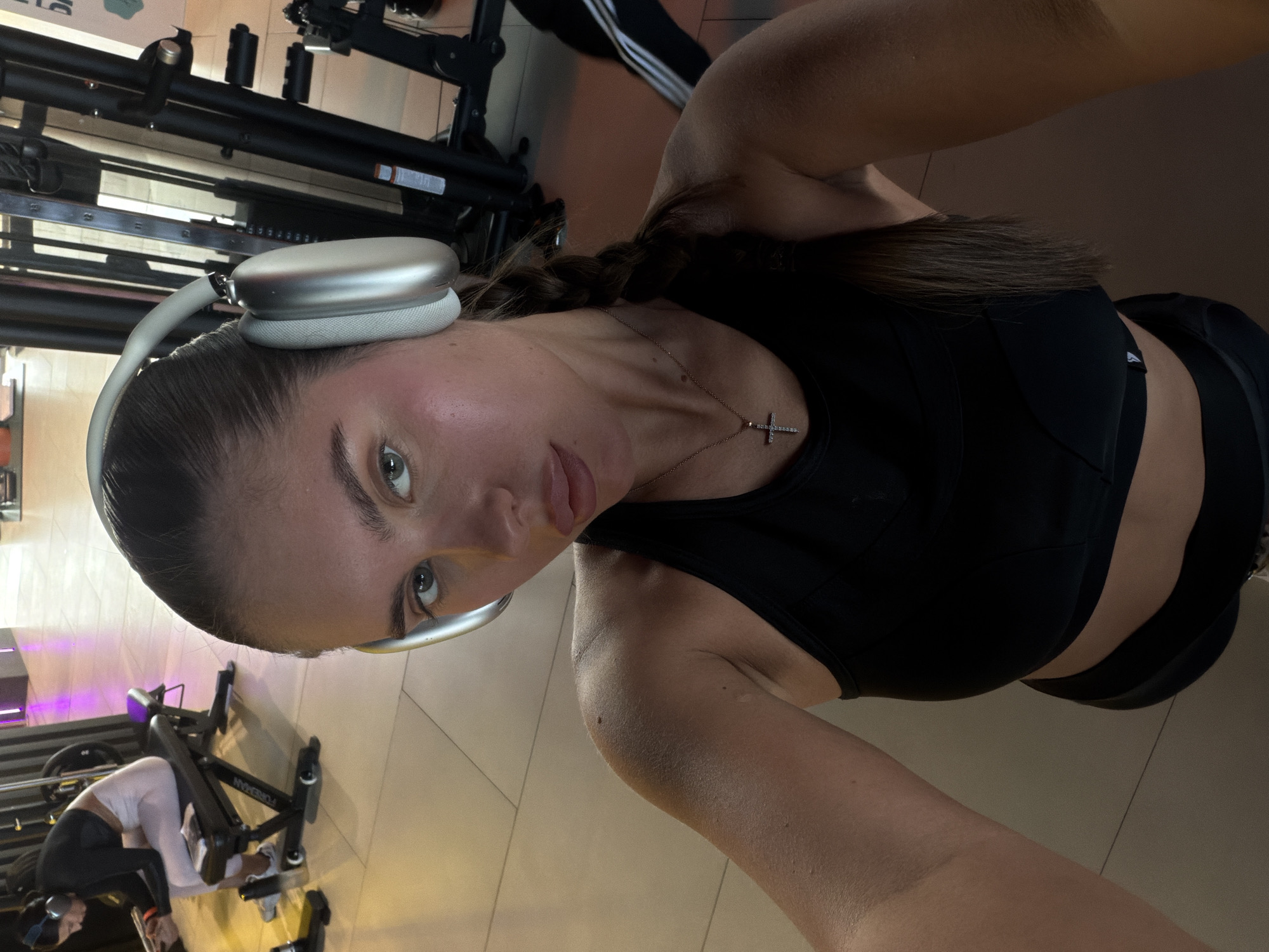 A woman with brown hair in a braid and wearing silver headphones is posing for a selfie in a gym. She is wearing a black sports bra and a silver necklace with a cross pendant. Exercise machines are visible in the background. The location is likely a gym in Krasnoyarsk, Russia.