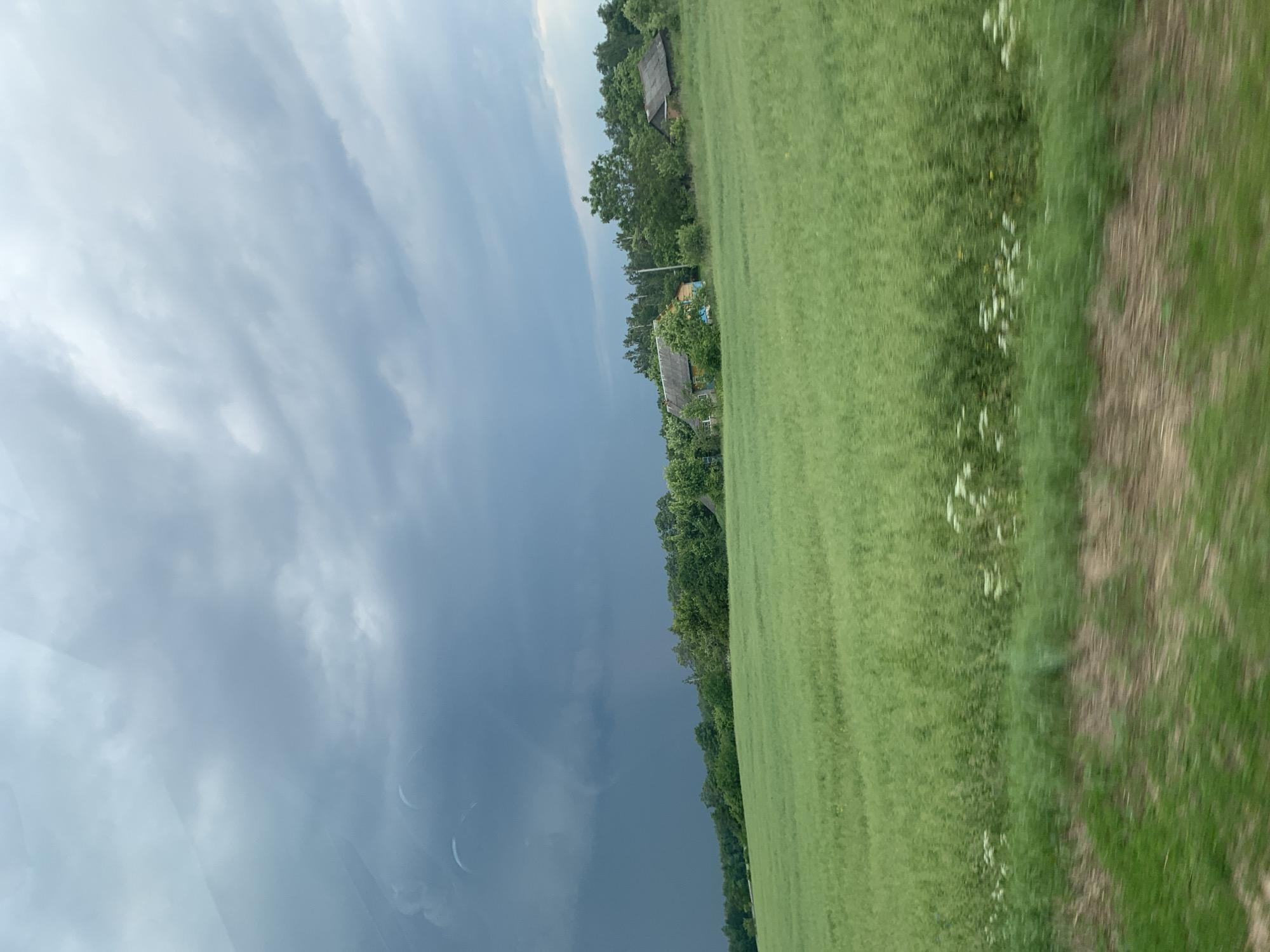 This media file depicts a rural landscape in Belarus, specifically near Слабада-Кучынскі сельскі Савет. The scene is divided diagonally, with a dramatic, dark, and cloudy sky dominating the left side, suggesting impending rain or a storm. The right side of the image features a vibrant green field, likely agricultural, with a dirt path running alongside it. Scattered white flowers are visible in the field.
Along the treeline bordering the field, several houses with grey roofs are nestled amongst lush green trees. These houses appear to be simple, rural dwellings. The time of day is difficult to ascertain precisely due to the overcast conditions, but the diffused light suggests it could be late afternoon or early evening.
The overall mood of the image is one of nature's power and tranquility, with the stark contrast between the stormy sky and the peaceful, green countryside. There is no visible text in the image. The perspective suggests the photo was taken from a moving vehicle, possibly a car, due to the slight motion blur in the foreground and the angle of the scene.