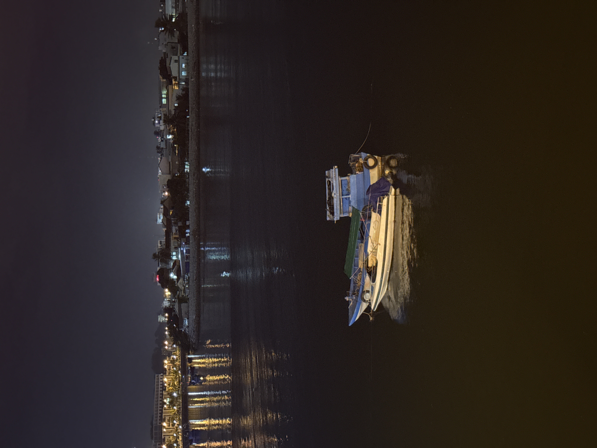 A nighttime scene captures a body of water with a city skyline in the background. Two boats are docked in the foreground, one larger and blue with white trim, the other smaller and white with blue trim. The larger boat has a covered section and a tire for cushioning. The water reflects the lights from the city, creating streaks of yellow and white. The city skyline features buildings with illuminated windows and streetlights, suggesting a populated area. The sky is dark and clear. The overall atmosphere is calm and serene, with the lights of the city providing a warm glow on the dark water. The location is likely near the Nha Trang River, given the cityscape and water.