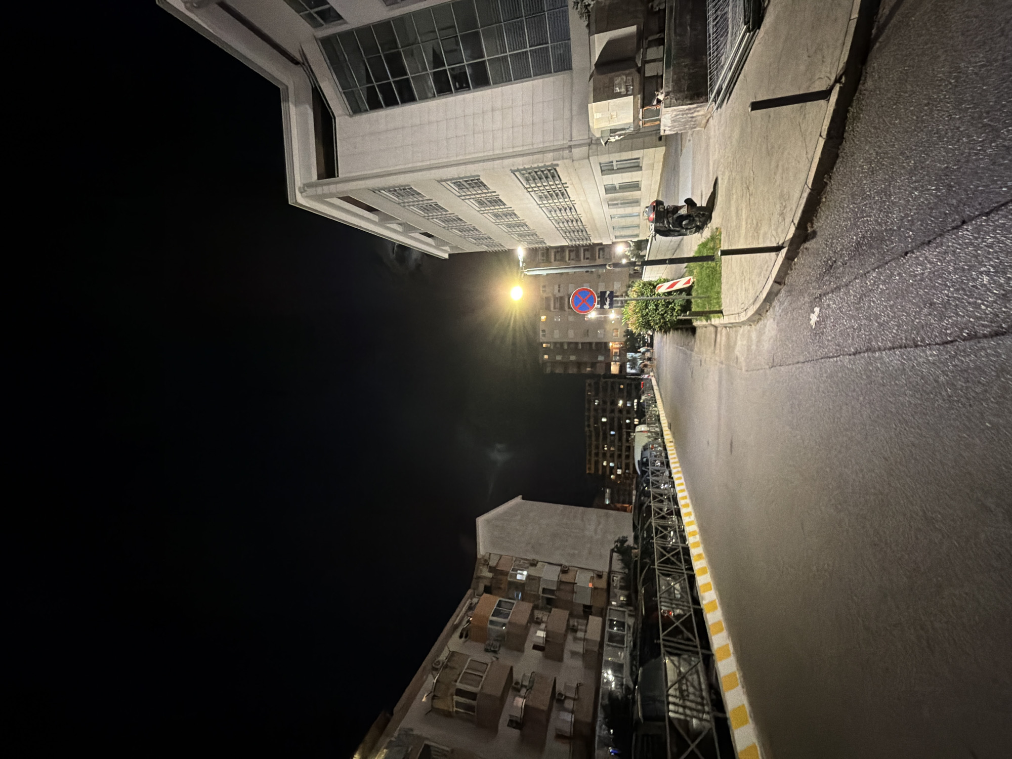 The image shows a street scene at night in Budva, Montenegro. On the left side of the street, a multi-story apartment building with balconies is visible, with parked cars lined up in front of it, separated by a yellow and white striped barrier. On the right side of the street, a modern white building with large windows dominates the scene. A street lamp illuminates the asphalt road and sidewalk. A red and blue circular traffic sign with a red line through it, indicating no parking or stopping, is present on the sidewalk. A parked motorcycle is also visible on the right side. Further down the street, other buildings with lit windows are discernible in the distance. The sky is dark with some faint cloud cover.