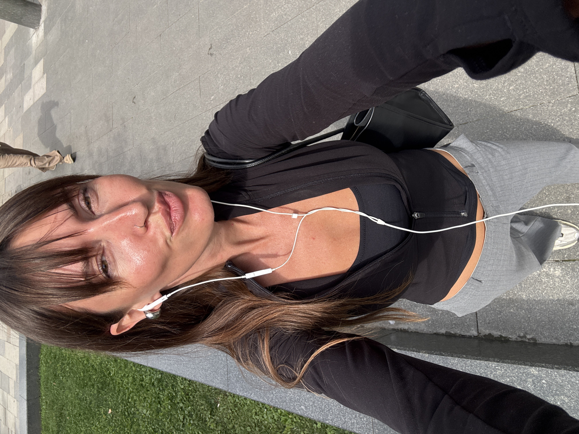 In a selfie taken outdoors, a woman with long brown hair and bangs, wearing white earbuds, a black zip-up jacket over a black sports bra, and grey pinstripe pants, poses with her lips pursed. She is standing on a paved area, possibly a sidewalk, with a grassy area to her left and a blurry figure walking in the background. The scene appears to be in an urban park or public space.