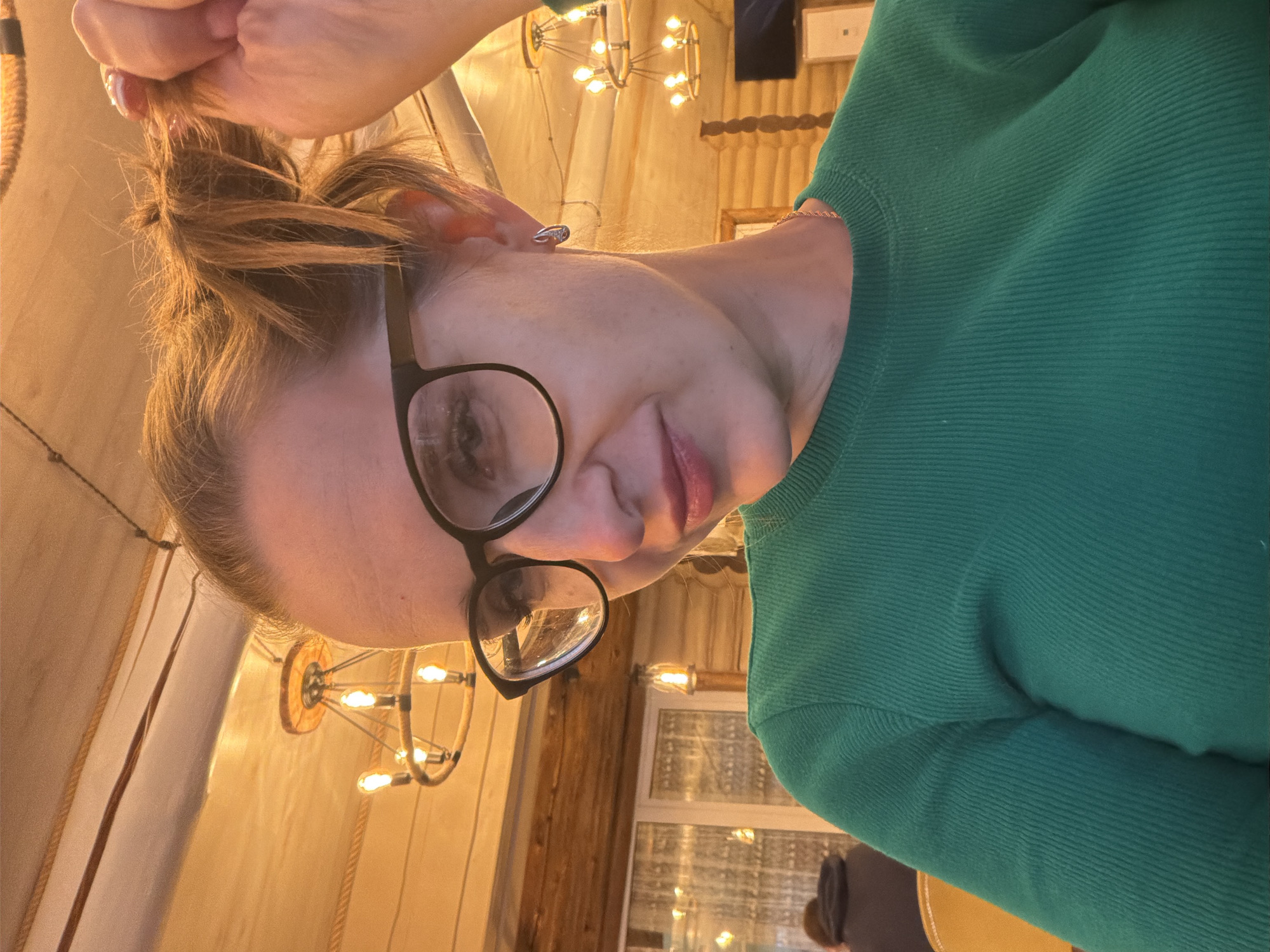 A woman wearing glasses and a green ribbed sweater is positioned in the foreground, partially holding her reddish-blonde hair. The background shows a room with chandeliers, suggesting a cafe or restaurant. A window and a wooden structure are also visible in the background. The location is indicated as Zaplav'e, Russia.