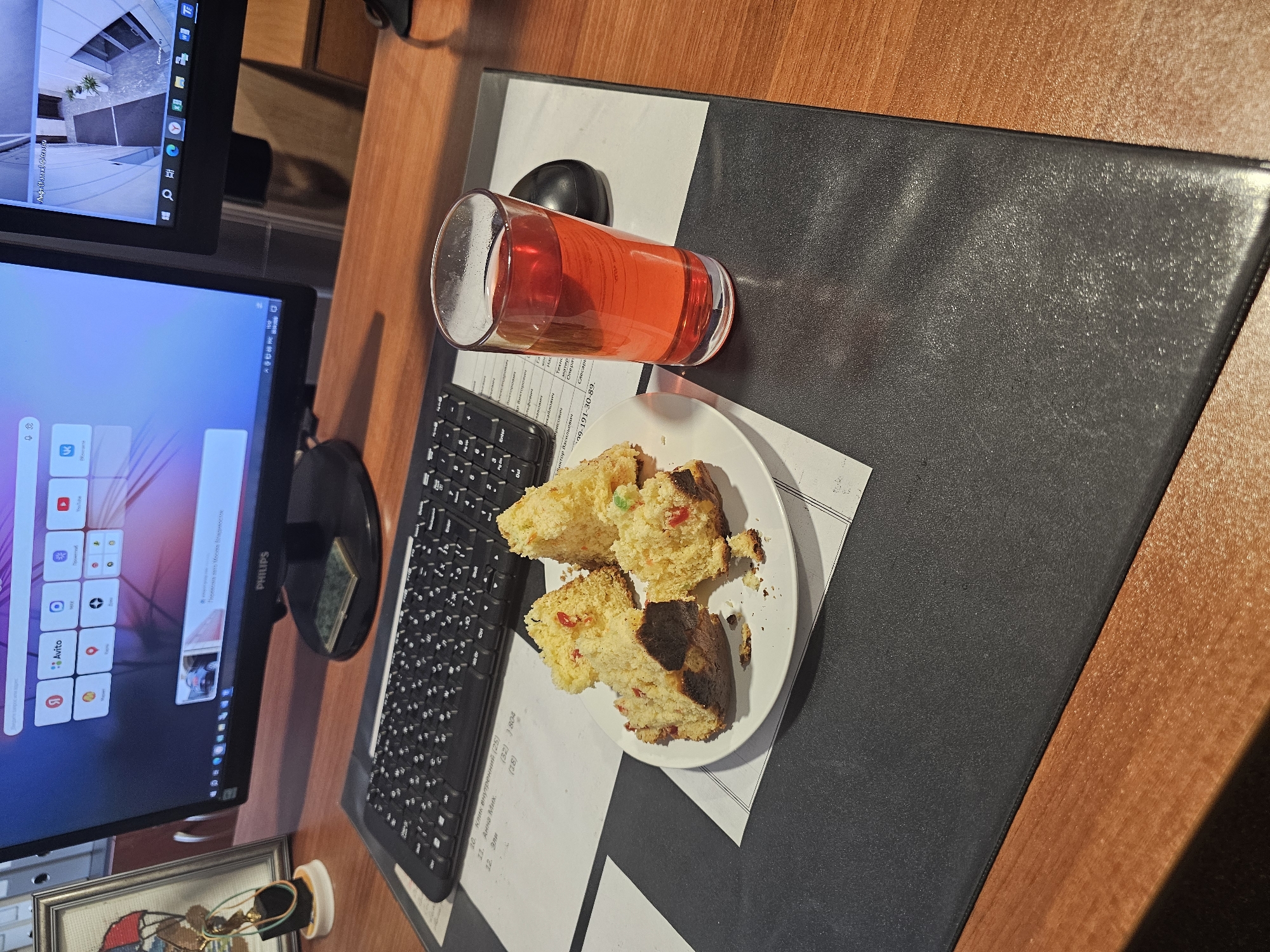 On a wooden desk, a monitor displays Russian text and icons. A glass of reddish liquid and a plate of cake sit on a black and white patterned mat next to a keyboard. Beneath the monitor, a framed item sits partially visible. OCR text on a paper indicates locations in Moscow, Russia.