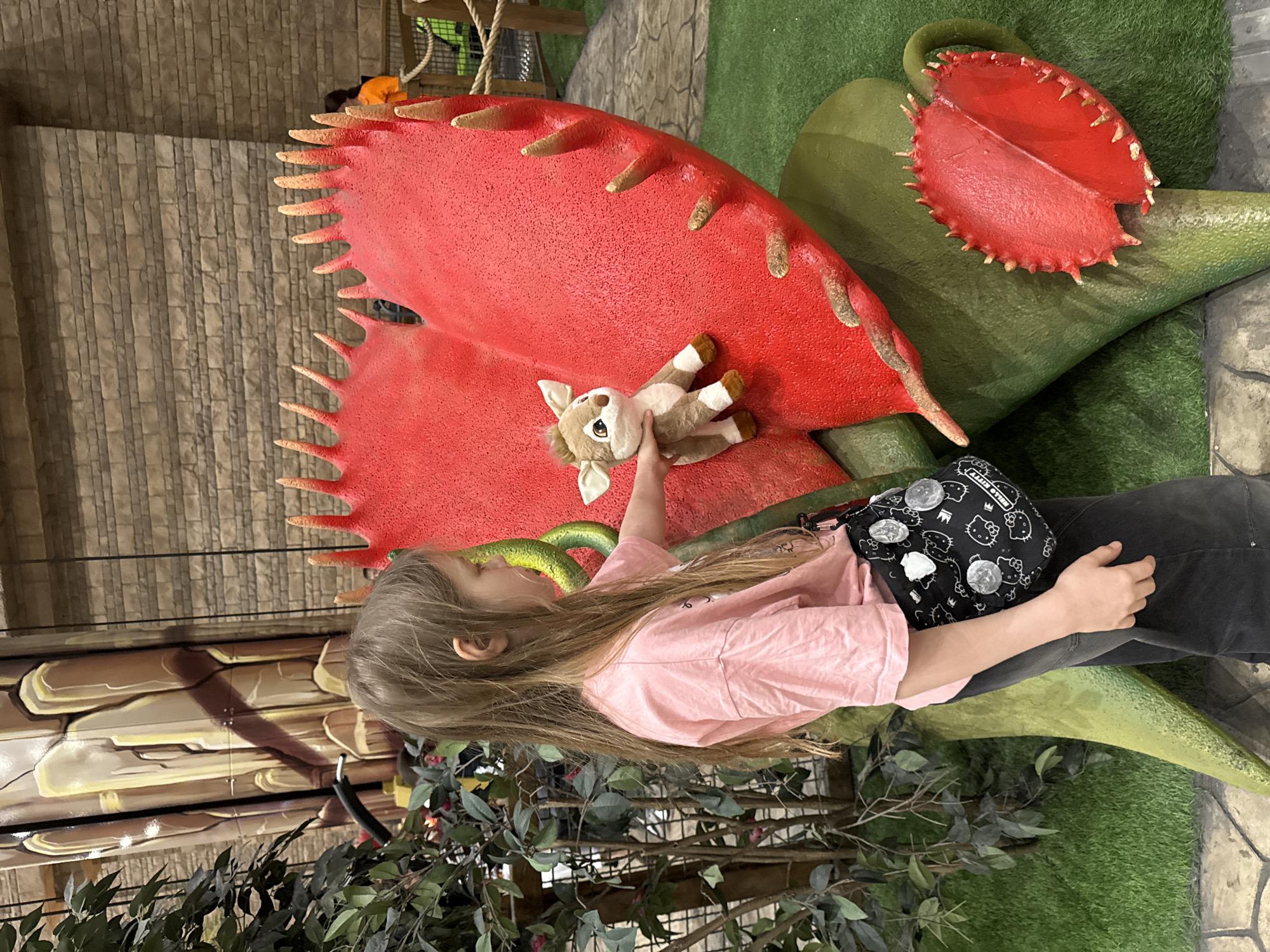 An indoor, eye-level shot captures a young girl interacting with a large, vibrant red model of a Venus flytrap within a themed exhibit in Moscow, Russia. The girl, with long, light brown hair, is seen from behind, wearing a light pink t-shirt and dark pants. She holds a small, light brown deer plush toy, playfully positioning it near the open "mouth" of the giant carnivorous plant model.
The flytrap model features prominent, pointed teeth-like structures along its red edges, and it emerges from a large, green, leaf-like base. Another, smaller red flytrap model is partially visible in the background to the right. The ground is covered with artificial green grass, and artificial dark green foliage frames the foreground and left side of the image.
The girl wears a black shoulder bag adorned with multiple white Hello Kitty faces and several silver pins. The text "HELLO KITTY" is clearly visible on the bag. The background features a textured wall resembling stacked stone or bricks, and hints of a rope structure suggest an indoor play area or museum exhibit. The lighting suggests an indoor setting with no specific time of day discernible.