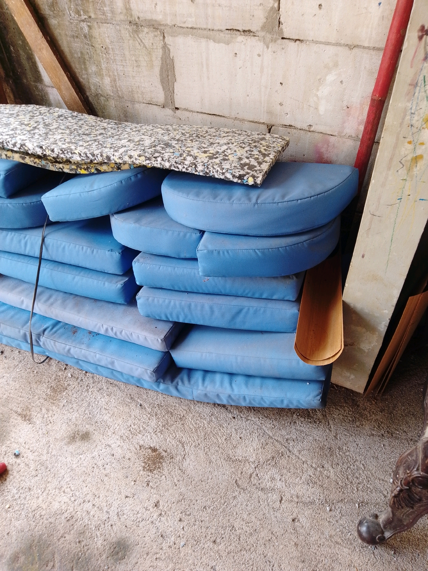 The image depicts a stack of blue cushions and a piece of foam padding leaning against a concrete block wall. To the right of the cushions, a red pole and a piece of light-colored material with paint splatters are visible. The scene appears to be an outdoor or workshop setting with a concrete floor. The location is identified as Cikal, Indonesia. There are no people or discernible interactions. The objects are static. The environment is somewhat cluttered and appears to be utilitarian.