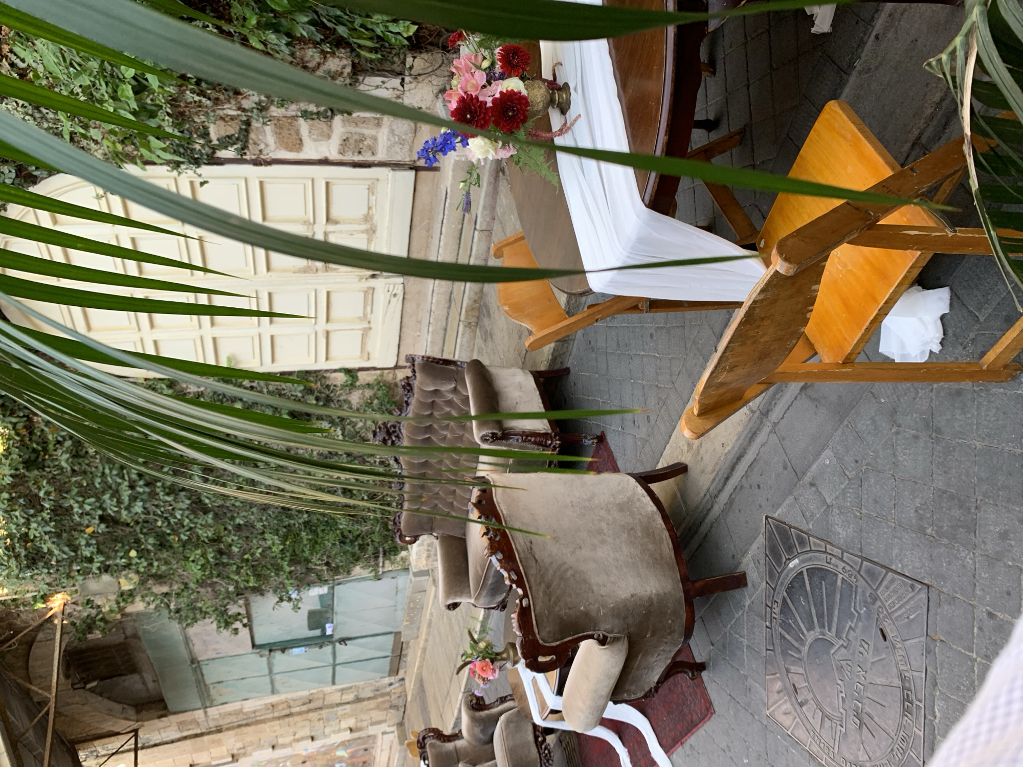 The image shows an outdoor seating area on a cobblestone street in Tel Aviv, Israel. In the foreground, several antique-style velvet armchairs and a sofa are arranged around a low wooden table. A white runner is draped across the table, which holds a vase of colorful flowers, including red dahlias, pink lilies, and blue delphiniums. To the right, a wooden folding chair sits in front of a round wooden table, also draped with a white runner. In the background, a large double door with ornate detailing is set into a stone wall covered in ivy. A metal grate with Hebrew writing is visible on the street in the lower left corner. The overall impression is of a charming, vintage-inspired outdoor event setup.