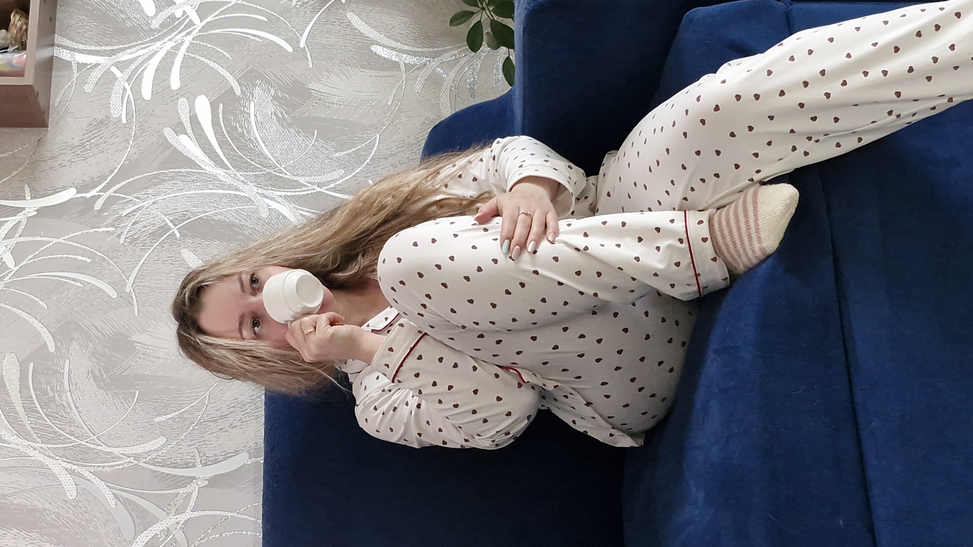 A woman with long blonde hair is sitting on a blue couch in what appears to be an indoor setting. She is wearing white pajamas with brown heart patterns and striped socks. She is holding a white cup to her face. The background includes a decorative wallpaper with a silver, swirling pattern. There is also a plant visible in the background. While it can't be determined with certainty if the image was taken in Otchor, Russia, there is nothing specific in the image to suggest that it was.