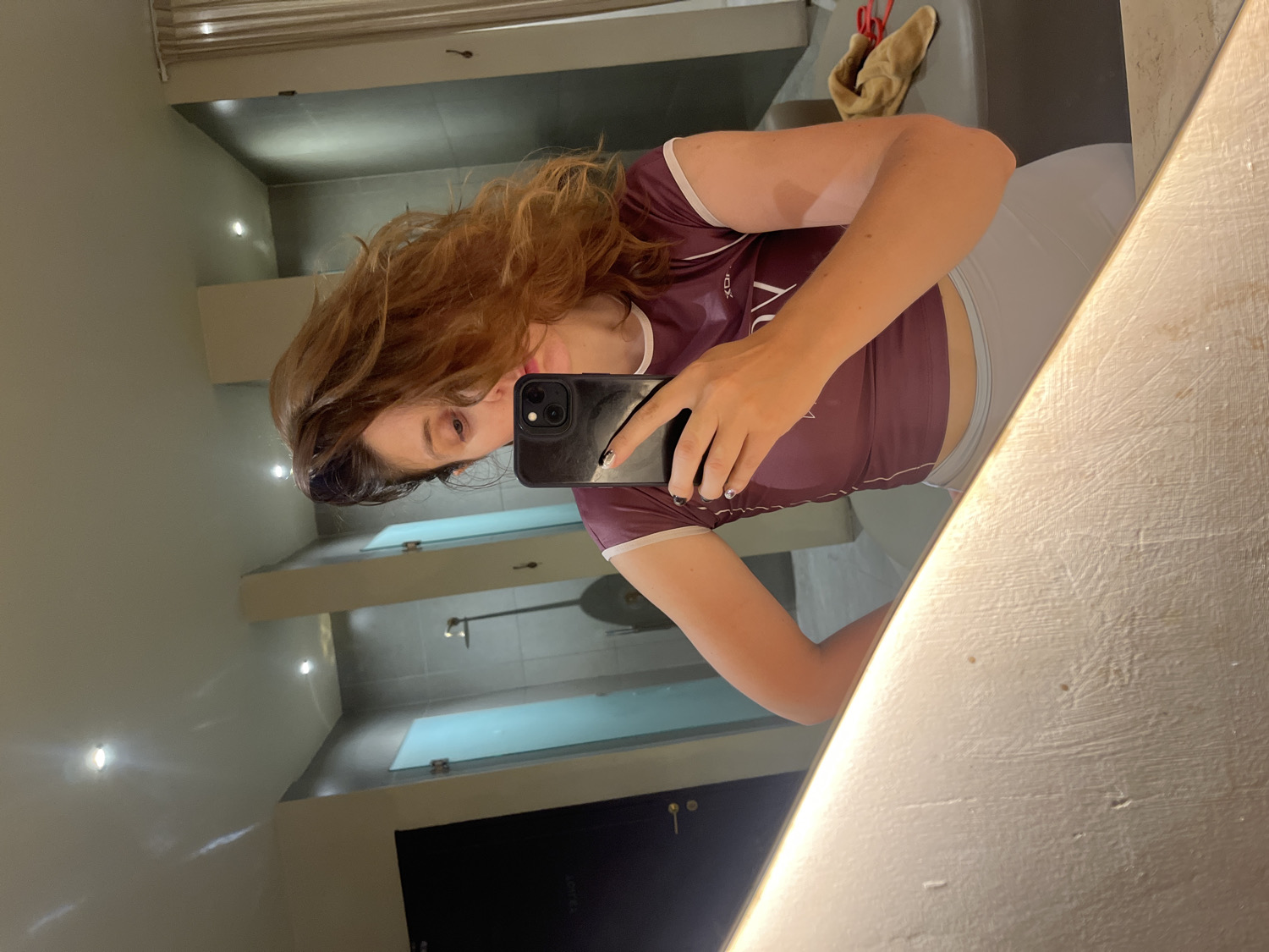 The image is a mirror selfie taken by a woman, rotated 90 degrees counter-clockwise. The subject is a young woman with long, wavy reddish-brown hair, prominently featured on the right side of the frame. She is holding a black smartphone with multiple cameras, likely an iPhone, in her right hand to capture her reflection. Her face is partially visible, showing her right eye, nose, and mouth with what appears to be light pink lipstick. She wears a maroon athletic top, possibly a rash guard, with light-colored trim on the sleeves. Visible white text on her chest reads "VOX". She is also wearing light grey or white athletic shorts or bottoms, visible around her waist. Her fingernails are painted a dark color.
The background, reflected in the mirror, depicts a modern indoor setting, likely a bathroom or changing room in a gym, spa, or hotel in North Kuta, Indonesia. The walls are light grey, and several overhead spotlights illuminate the space. In the upper left corner of the reflection, a light brown towel is draped over a fixture. Further in the background, rectangular panels or doors, possibly for shower stalls or lockers, are visible. The vanity or counter surface where the mirror is mounted is a light, textured material, illuminated from one side. The reflection on her phone screen shows some inverted and unidentifiable text. The woman's expression is neutral, focused on the act of taking the photograph.