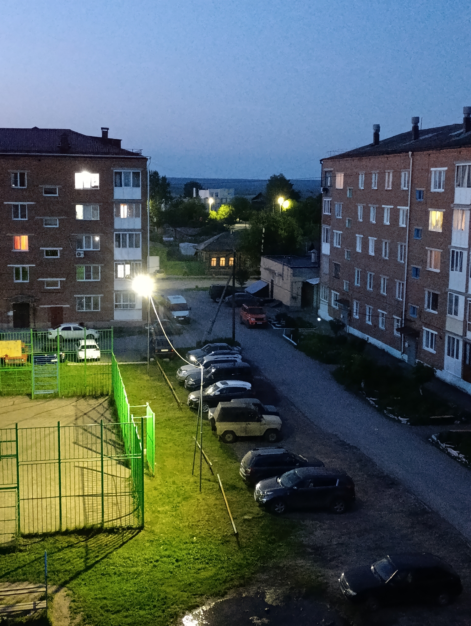 The image captures an evening view of a residential area in Tara, Russia, from an elevated perspective, likely a window or balcony. The scene is dominated by two multi-story brick apartment buildings, one on the left and another on the right, typical of post-Soviet architecture. Many windows in both buildings are illuminated, suggesting residents are home.
In the foreground, to the left, there is a fenced sports court, possibly a basketball or soccer pitch, with a green metal fence and a brightly lit surface, although it appears empty. A cluster of parked cars, including a white sedan and a white hatchback, are visible near the building on the left.
A paved road or driveway runs between the two main buildings, lined with more parked vehicles, predominantly dark-colored SUVs and sedans. A red car, possibly an older SUV model, is distinctive in the middle ground. Bright streetlights cast strong, yellowish-green light on the ground, creating distinct shadows and illuminating the grassy areas and the road. The sky above is a deep twilight blue, indicating it is late evening or early night.
The overall atmosphere is quiet and still, with no human activity directly visible in the frame, but the lit windows and numerous parked cars convey a sense of a populated, residential environment. Trees and distant buildings are faintly visible under the dark sky towards the horizon. There is no visible text in the image.