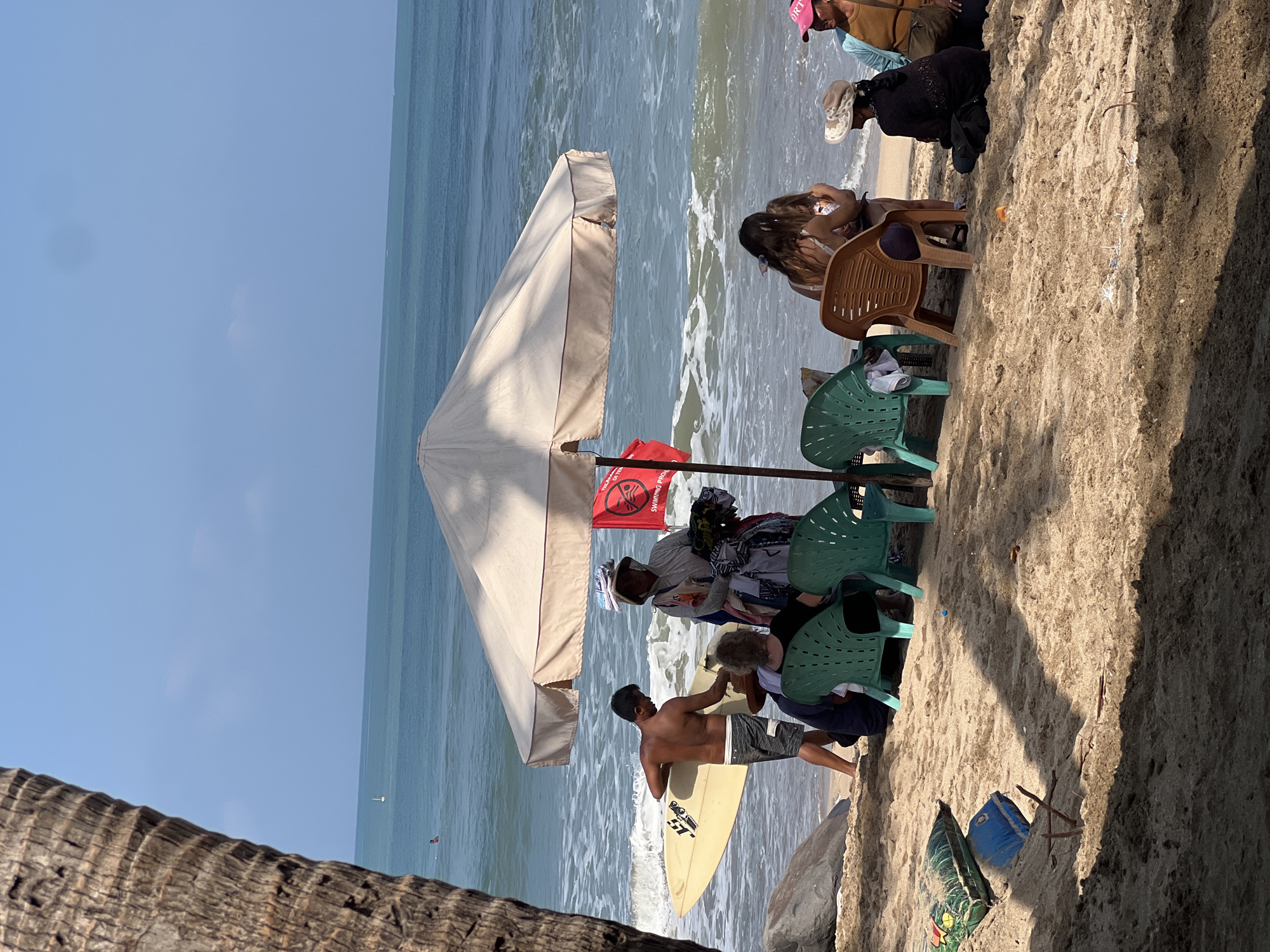 A group of people are on a beach in Kuta, Indonesia. The ocean is visible in the background with waves breaking on the shore. A man holding a surfboard is standing near the water, and a vendor is selling items under a large umbrella. Several people are sitting on chairs on the sand, some in front of the vendor and some further away. The weather appears to be sunny with a clear blue sky. The sand is light brown and textured, with some shadows cast by the umbrella and palm trees.