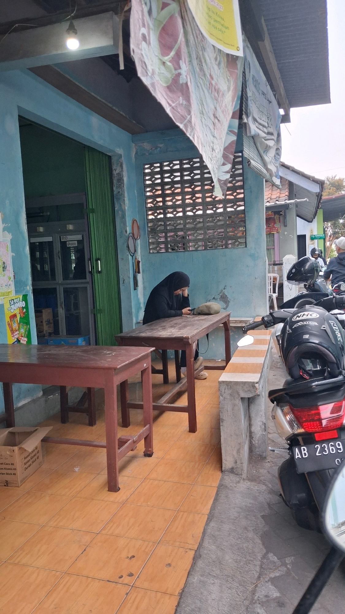 The image depicts an outdoor scene in front of a light blue building, likely in Yogyakarta, Indonesia, given the context. A woman wearing a black hijab is seated at a wooden table, looking down at her phone. She is positioned on the right side of the frame. In the foreground, two wooden tables are visible. The table on the left has a cardboard box on it, and the table on the right is where the woman is seated. The building features a green folding door to the left, and a window with a decorative grate. Two badminton rackets are hanging on the wall above the woman. A portion of a motorcycle with the license plate "AB 2369" is visible on the right side of the image, along with a black helmet. The ground is tiled with orange square tiles. The overall impression is of a simple, possibly local establishment.