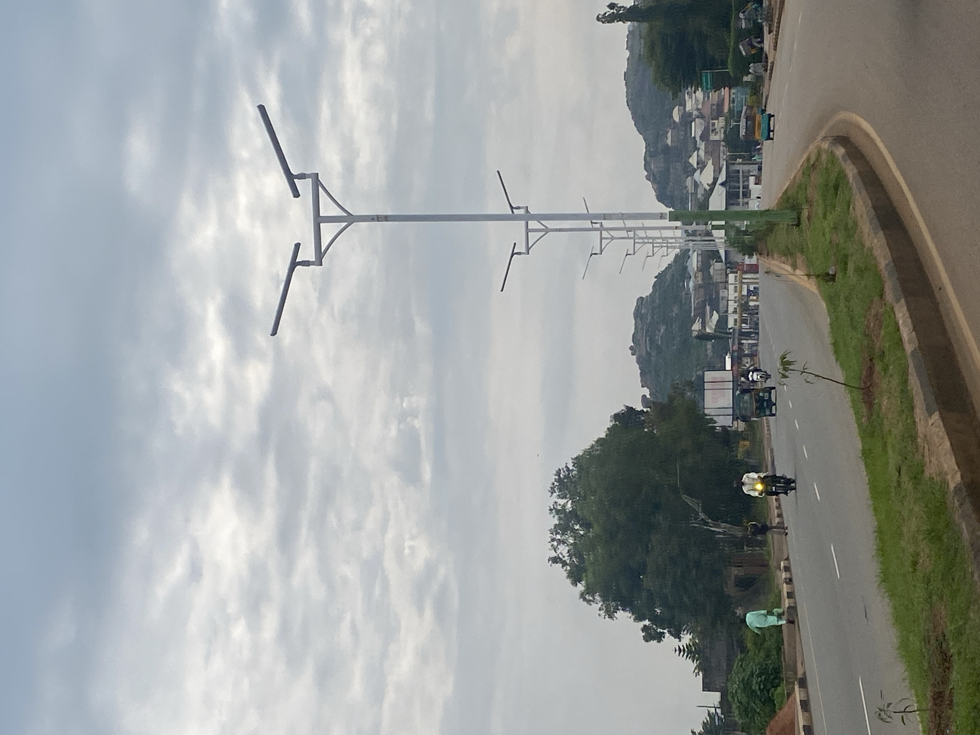 The image depicts a view of a road in the city of Jos, Nigeria, under a cloudy sky. The road is paved with asphalt and has multiple lanes. Several streetlights with multiple arms line the side of the road. A large green tree stands prominently on the left side of the road, with a residential area and rocky hills visible in the background. On the road, a person in a light blue garment is walking, while another person rides a motorcycle. Further down the road, an auto-rickshaw is seen, followed by another motorcycle. The environment appears to be a mix of urban and natural landscapes, with buildings nestled among the hills. The overall impression is of a typical day in a Nigerian city.