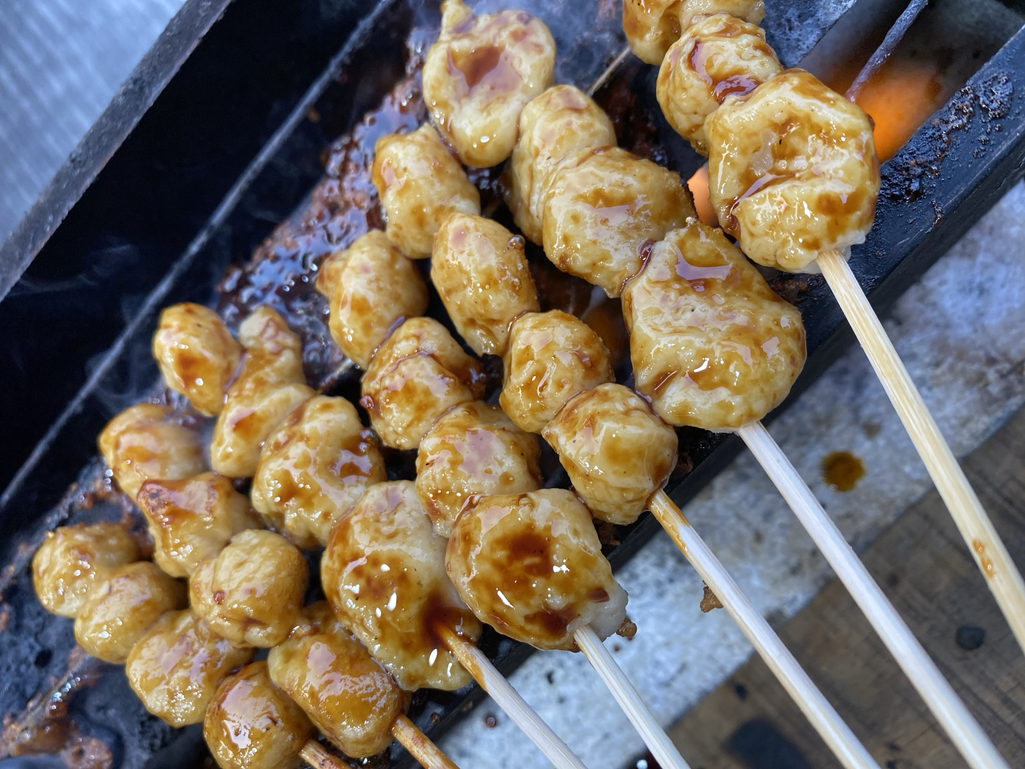 A close-up, high-angle shot showcases several skewers of sweet and savory glazed meatballs being grilled over an open flame. The meatballs are round and plump, coated in a shiny, translucent glaze that has caramelised in places. They are skewered onto thin, light-colored wooden sticks, with multiple meatballs per skewer. The grilling surface is a black, slatted metal grate, beneath which a bright orange flame is visible. The background is dark and out of focus, with the focus sharply on the food. The scene appears to be outdoors, possibly at a street food stall in Kasihan, Indonesia, given the style of food and grilling method.