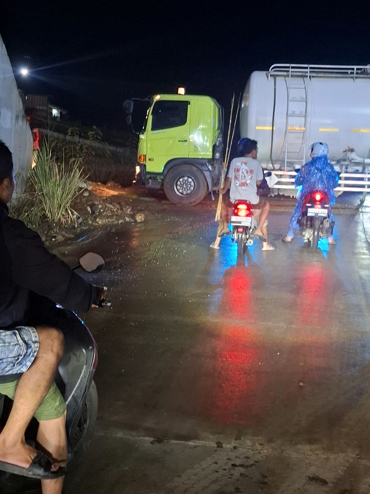 At night in Ciheulang Tonggoh, Indonesia, a lime green truck and a white tanker truck are positioned on a wet, muddy road. Two motorcycles are stopped behind the green truck. The motorcycle on the left has two individuals; the driver is wearing a black jacket and denim shorts, and a second person is seated behind them. The motorcycle on the right has one individual wearing a blue rain suit and a blue helmet. The rear brake lights of both motorcycles are illuminated, casting red reflections on the wet road surface. The environment appears to be a rural or semi-rural area, with some vegetation visible to the left of the road. The ground is uneven and shows signs of recent rainfall.