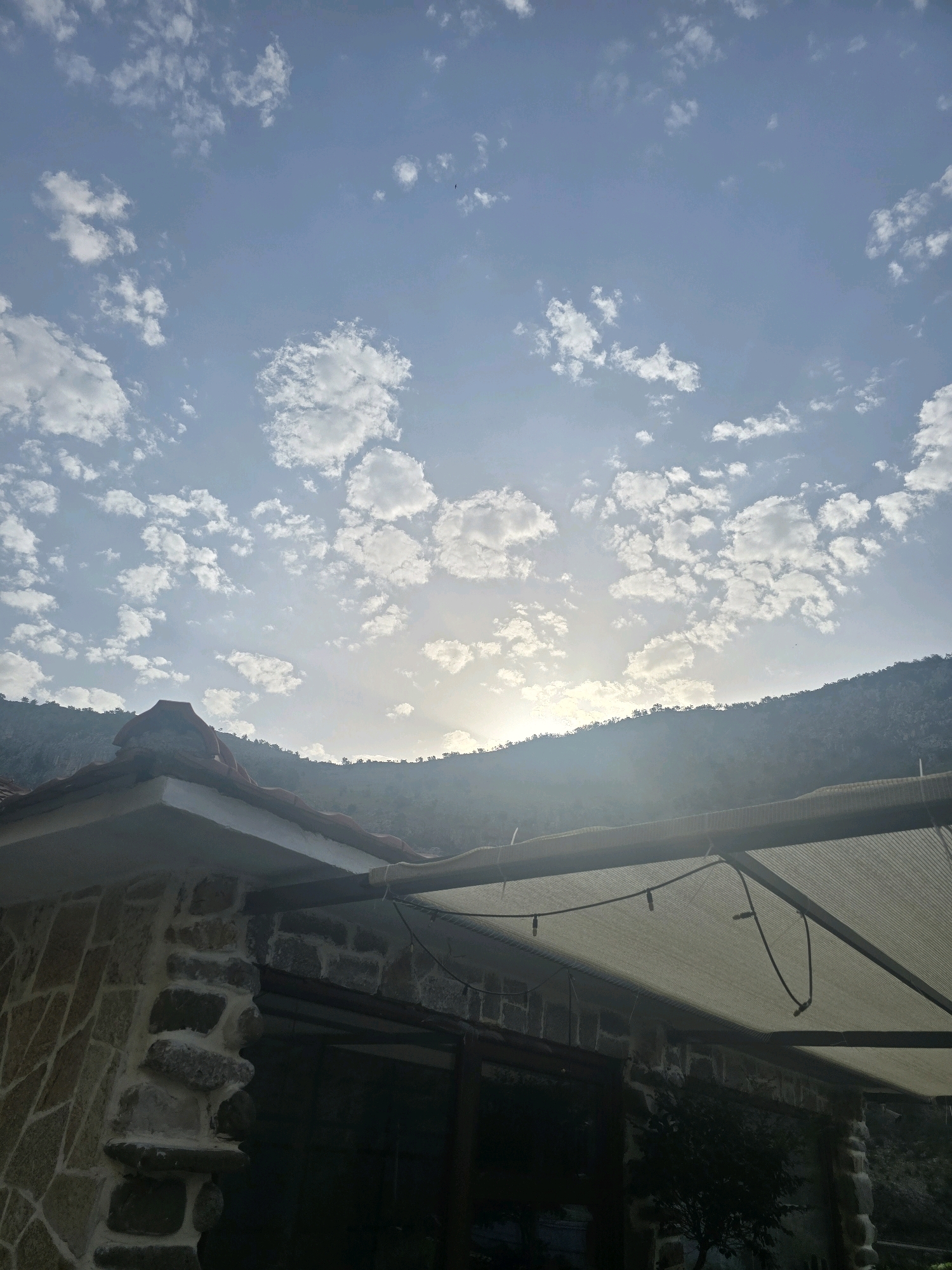 The image captures a scenic view of a mountainous landscape with a building in the foreground. The sky is a light blue, scattered with fluffy white clouds, and the sun is partially visible, creating a soft glow. A mountain range, covered in dense vegetation, forms the horizon line. In the lower left of the frame, a building with a stone facade and a red-tiled roof is visible. A covered patio area with a light-colored fabric awning extends from the building. The image appears to be taken from an outdoor perspective, possibly a residential area or a tourist spot. The location is identified as Podgorica, Montenegro.