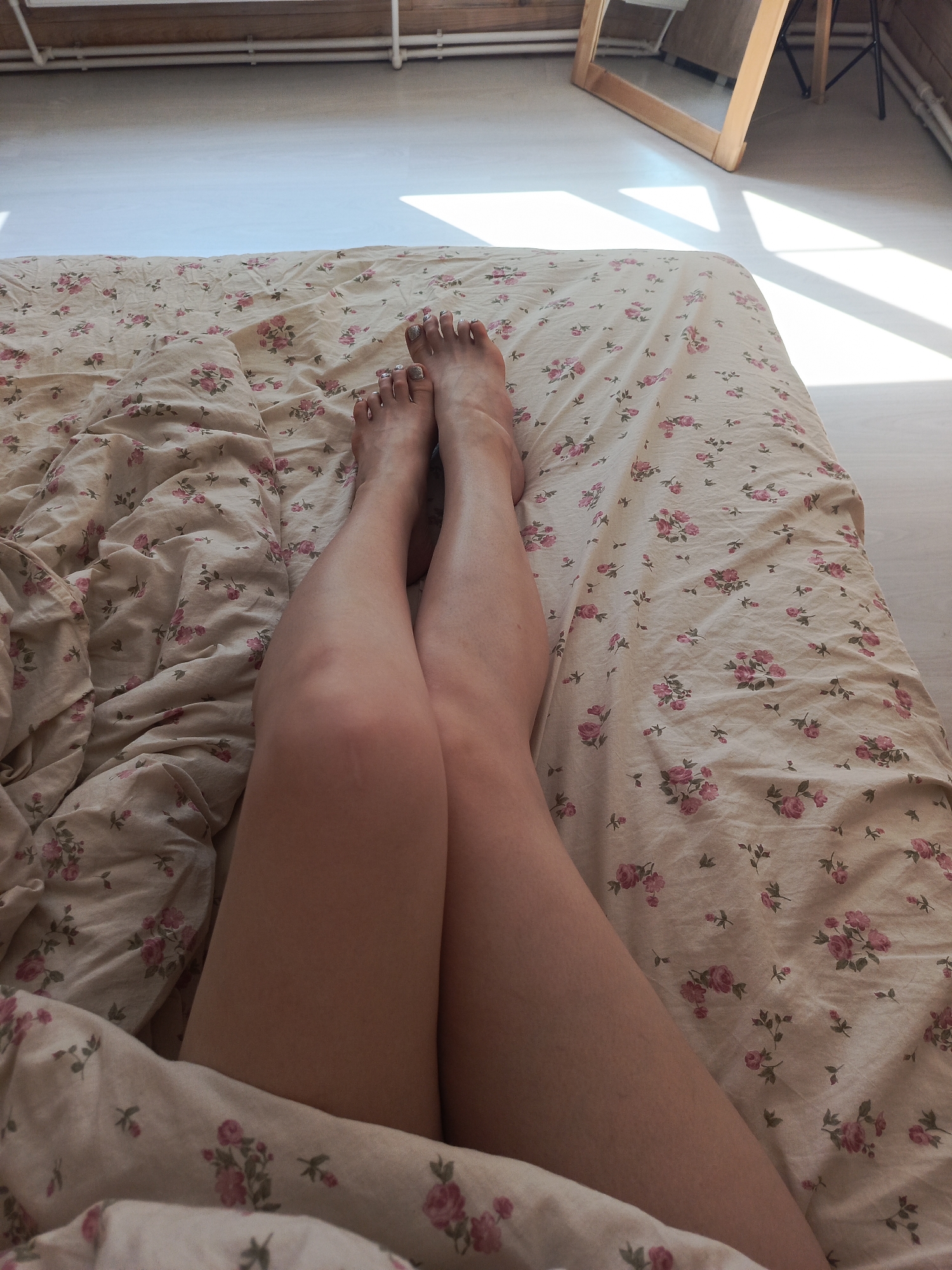 The image depicts a close-up view of a person's legs and feet resting on a bed with a floral-patterned bedsheet. The person is lying down, with their legs crossed at the knees. The background reveals a room with light-colored walls and a wooden floor. A mirror is leaning against the wall in the background. The location is Северный, Russia.