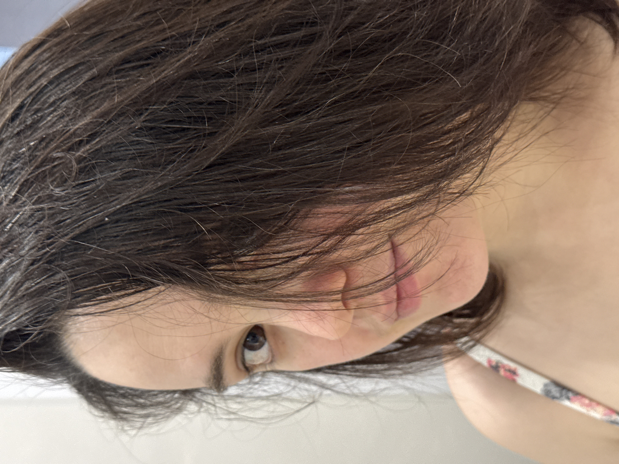 The image displays a close-up, chest-up portrait of a female individual. She possesses fair skin and dark brown, slightly disheveled hair, which falls across and partially obscures the right side of her face. Her left eye, featuring a dark brown iris, is clearly visible and directed forward. The left side of her nose and her lips are partially visible. The individual is wearing a garment with a white, floral-patterned strap (red and blue hues) visible over her left shoulder. The background is a plain, light-colored wall or interior surface. No other subjects, objects, or actions are discernible. The specific location within Izhevsk, Russia, or any other city, cannot be determined from the visual information provided.