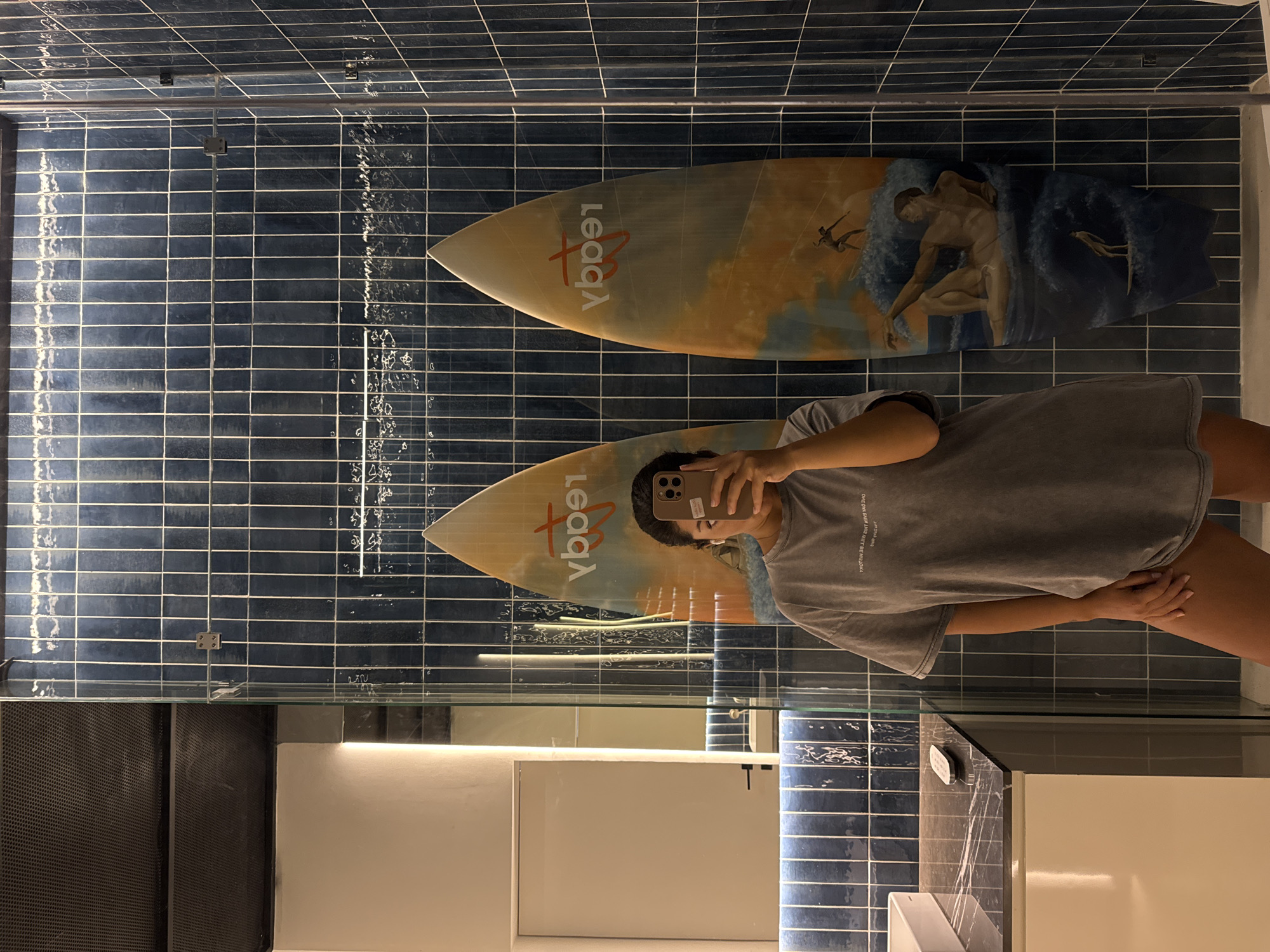 A woman is seen in a bathroom mirror selfie. She is holding a smartphone in front of her face. The bathroom features blue, tiled walls and two surfboards mounted above the sink. One surfboard displays an image of a nude man, perhaps an artistic depiction, while the other features the text "Legacy." There is a sink in the mirror.
This location is in Kuta Selatan, Indonesia.