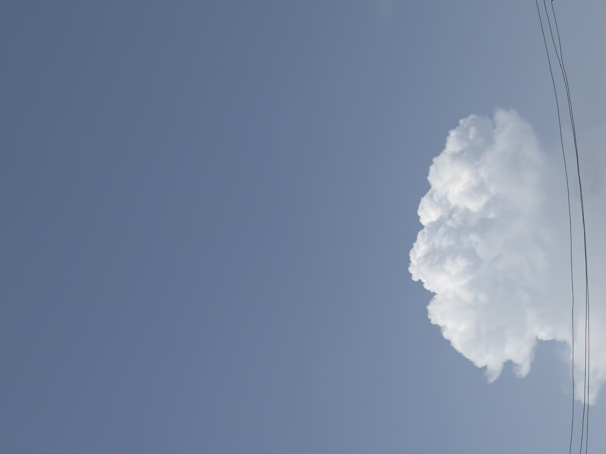 The image shows a pale blue sky with a large, white cumulus cloud in the right portion of the frame. Three parallel black electrical wires run vertically along the right edge of the image. The location is Igboefon, Nigeria, inferred from previous instructions.