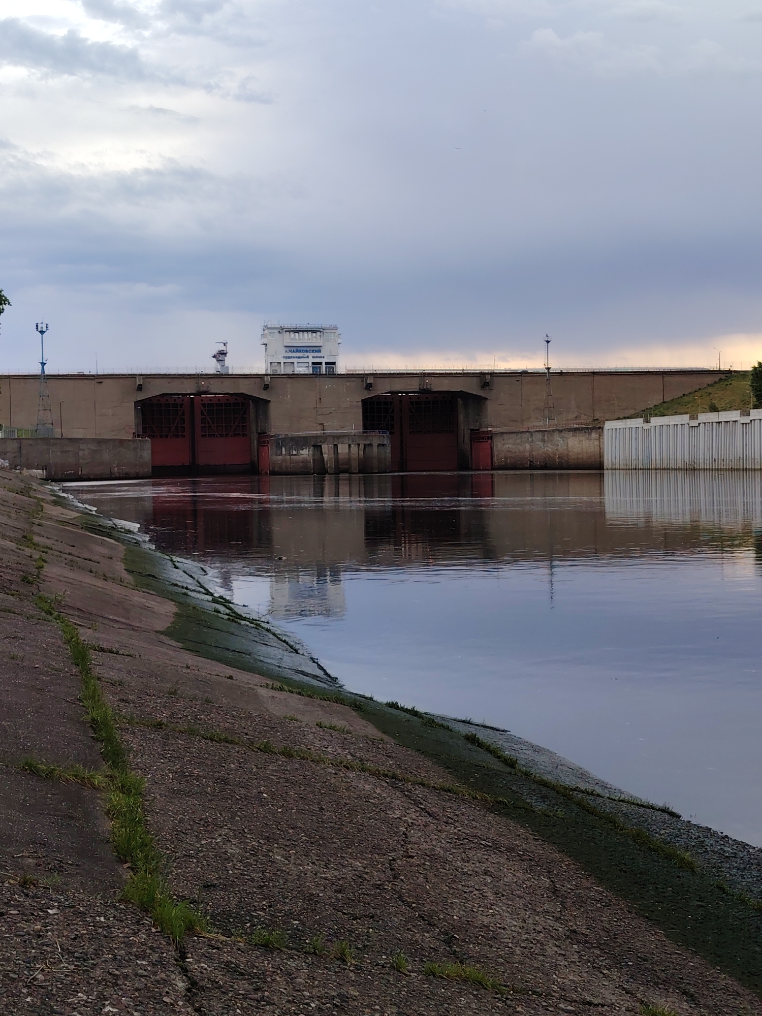 The image depicts a water scene, likely a river or reservoir, near a concrete embankment in the city of Chaikovsky, Russia. A dam or lock structure spans the waterway, featuring large red gates. A building, labeled in Cyrillic, is positioned atop the structure, possibly for control or administration. The water is still and reflects the dam and sky, which is overcast with heavy clouds. Vegetation is sparse, clinging to cracks in the concrete embankment.