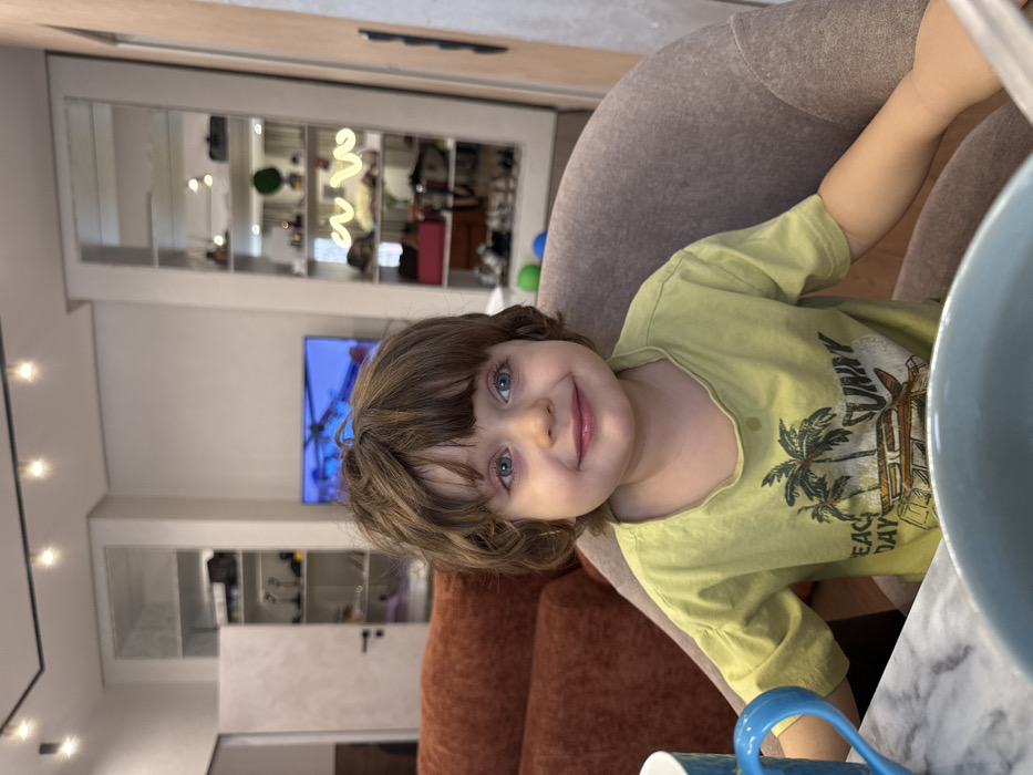 The image depicts a young child seated at a table in an indoor residential setting in Khimki, Russia. The child, with light brown, wavy hair and blue-green eyes, presents a smiling expression while looking directly at the camera. They wear a light green short-sleeved t-shirt adorned with a 'BEACH DAY SUNNY' graphic featuring palm trees and a vehicle; a small wet stain is visible on the front. The child occupies a light brown upholstered chair, situated adjacent to a visible section of an orange-brown upholstered sofa on the left. In the immediate foreground, a white and grey marble-patterned tabletop holds a blue textured mug and a partially visible light blue bowl. The background reveals a contemporary interior: a white ceiling with a black track lighting system, recessed shelving units with mirrored or glass fronts displaying various objects on both the left and right sides of the frame, and a wall-mounted flat-screen television displaying a colorful image. A white door is visible on the far left, and a light wooden door frame appears on the far right. One of the shelving units features a white wavy neon sign.
