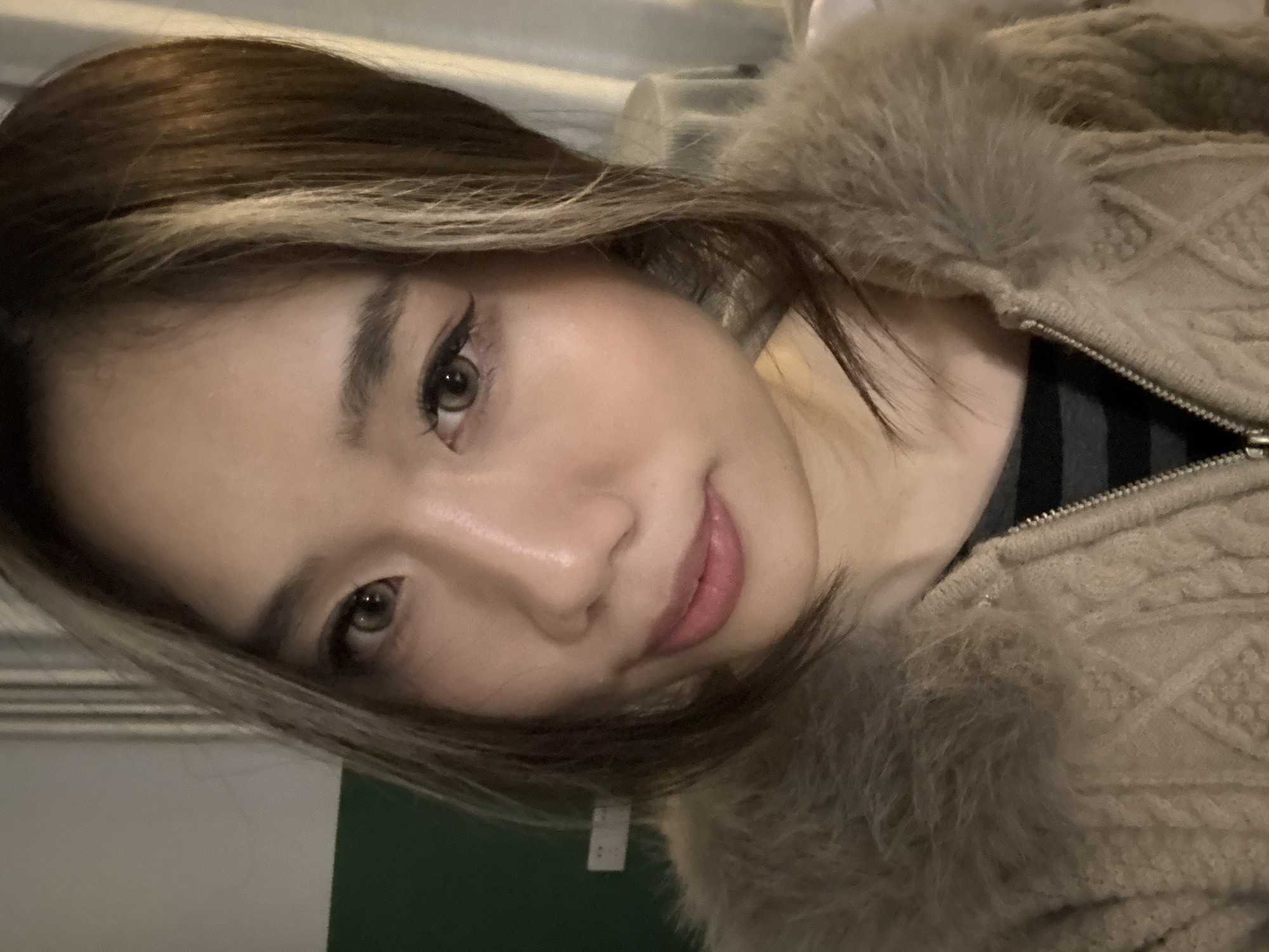 A close-up, waist-up portrait captures an East Asian female in an indoor setting. The individual occupies the majority of the frame, facing directly towards the camera with a slight, closed-lip smile. She has dark brown hair with distinct light brown or blonde streaks framing her face on the viewer's left. Her eyes are light brown, possibly enhanced by contact lenses, with visible black winged eyeliner and defined eyebrows. Her lips are full and coated with a light pink gloss. She is wearing a light brown or beige knitted jacket with a cable knit pattern and a prominent faux fur collar of a similar color. A silver-toned zipper is visible down the front. Beneath the jacket, a dark garment with horizontal stripes is partially visible. The background, in an unidentifiable indoor location, presents a wall section on the left with a lighter upper half and a dark green lower half, where a white rectangular object (possibly an outlet or switch) is mounted. The right side of the background shows indistinct, lighter-toned elements. The specific location in the city of Liangjiang, China, cannot be confirmed from the visual information provided within the image.