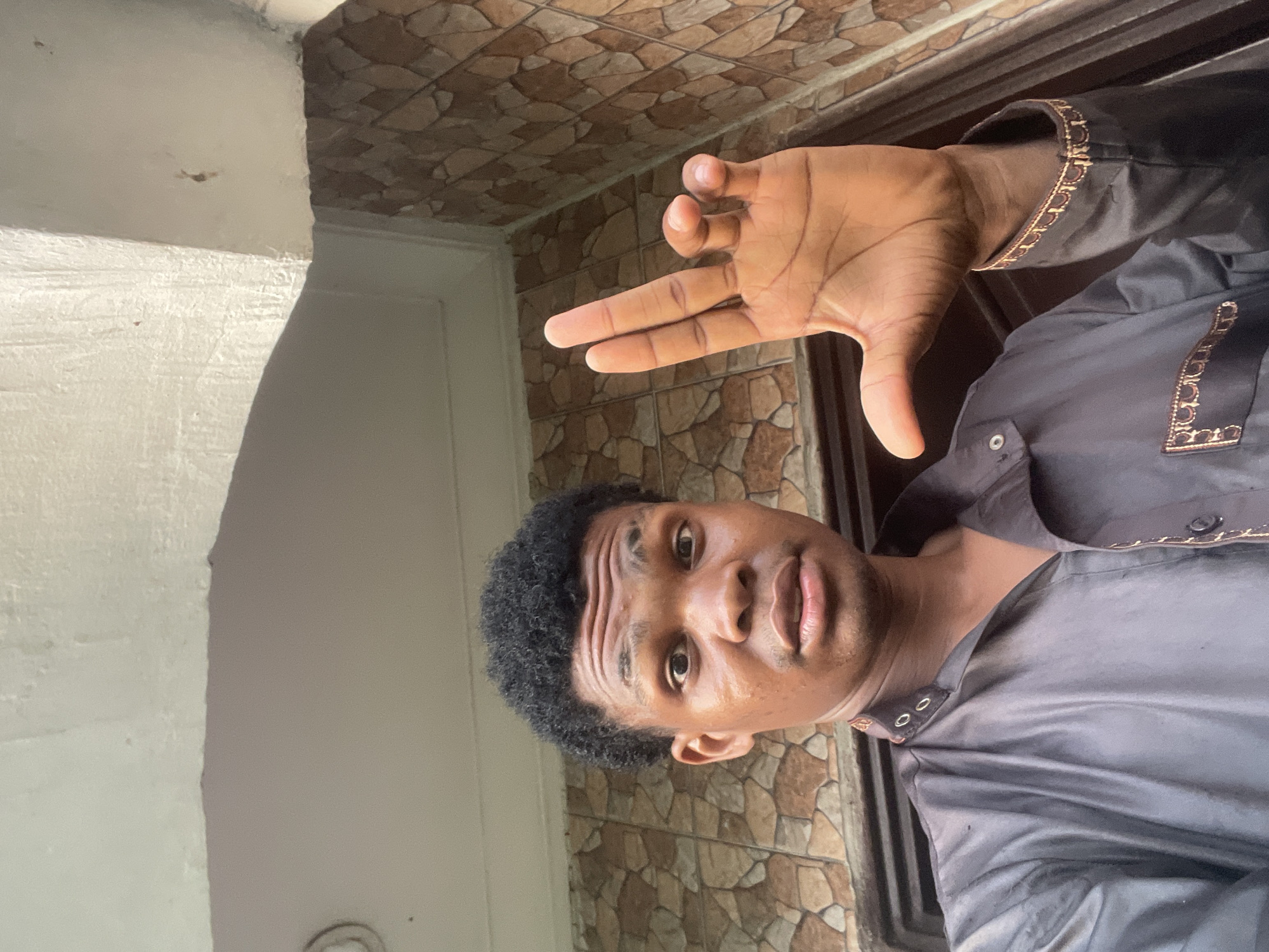 In Ado, Nigeria, a young Black male with dark skin and curly black hair is captured in a selfie. He is wearing a dark grey, possibly silk, button-down shirt with ornate gold trim around the collar and cuffs, and a pocket on the left side of his chest. His left hand is raised, displaying a gesture where his index and middle fingers are extended, and his ring and pinky fingers are curled downwards. The background consists of a textured, light brown wall, and an overhang of a white, textured ceiling. The lighting suggests an outdoor or semi-outdoor setting, possibly in front of a building entrance.