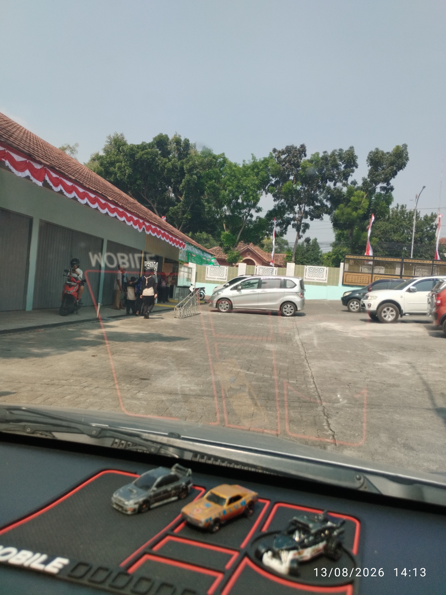 The image shows a street scene in front of a building decorated with red and white banners. Several cars are parked in the lot. In the foreground, on the dashboard of a vehicle, are three toy cars and a date stamp indicating "13/08/2026 14:13". The building appears to be a commercial or public facility. The overall atmosphere is casual and sunny, suggesting a daytime activity. There are trees and a clear sky in the background. The location is identified as Bandar Lampung, Indonesia.