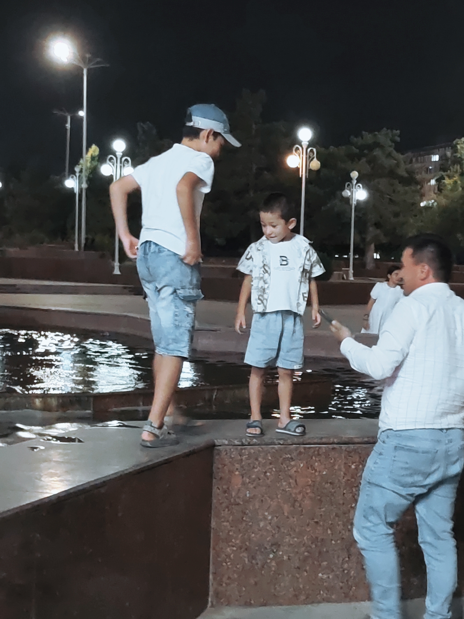 The image captures a nighttime scene in a public park in Tashkent, Uzbekistan. Three individuals are visible. In the foreground, a man wearing a white checkered shirt and light blue jeans holds a dark smartphone, seemingly interacting with a young boy. The boy, dressed in a white t-shirt with a patterned overshirt and light blue shorts, stands facing the man. Behind him, another boy, wearing a white t-shirt and light blue cargo shorts, stands near a body of water. The park features decorative lampposts and lush trees, illuminated by the artificial lights. The water reflects the surrounding lights, creating a shimmering effect. The overall atmosphere appears casual and relaxed, with the figures seemingly engaged in a pleasant evening outing.